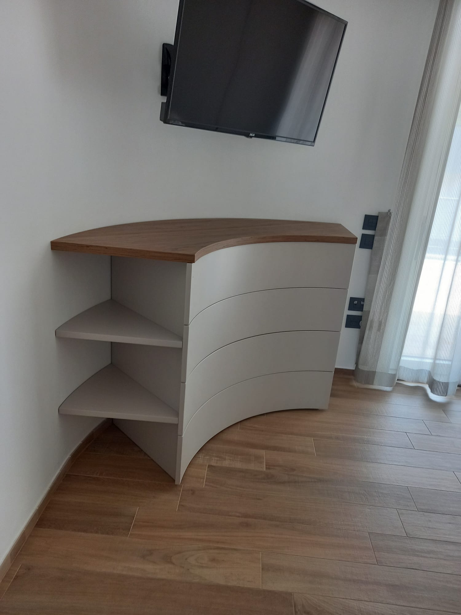 Un mobile bianco curvo con piano di lavoro e ripiani in legno, sotto una TV montata su una parete bianca.