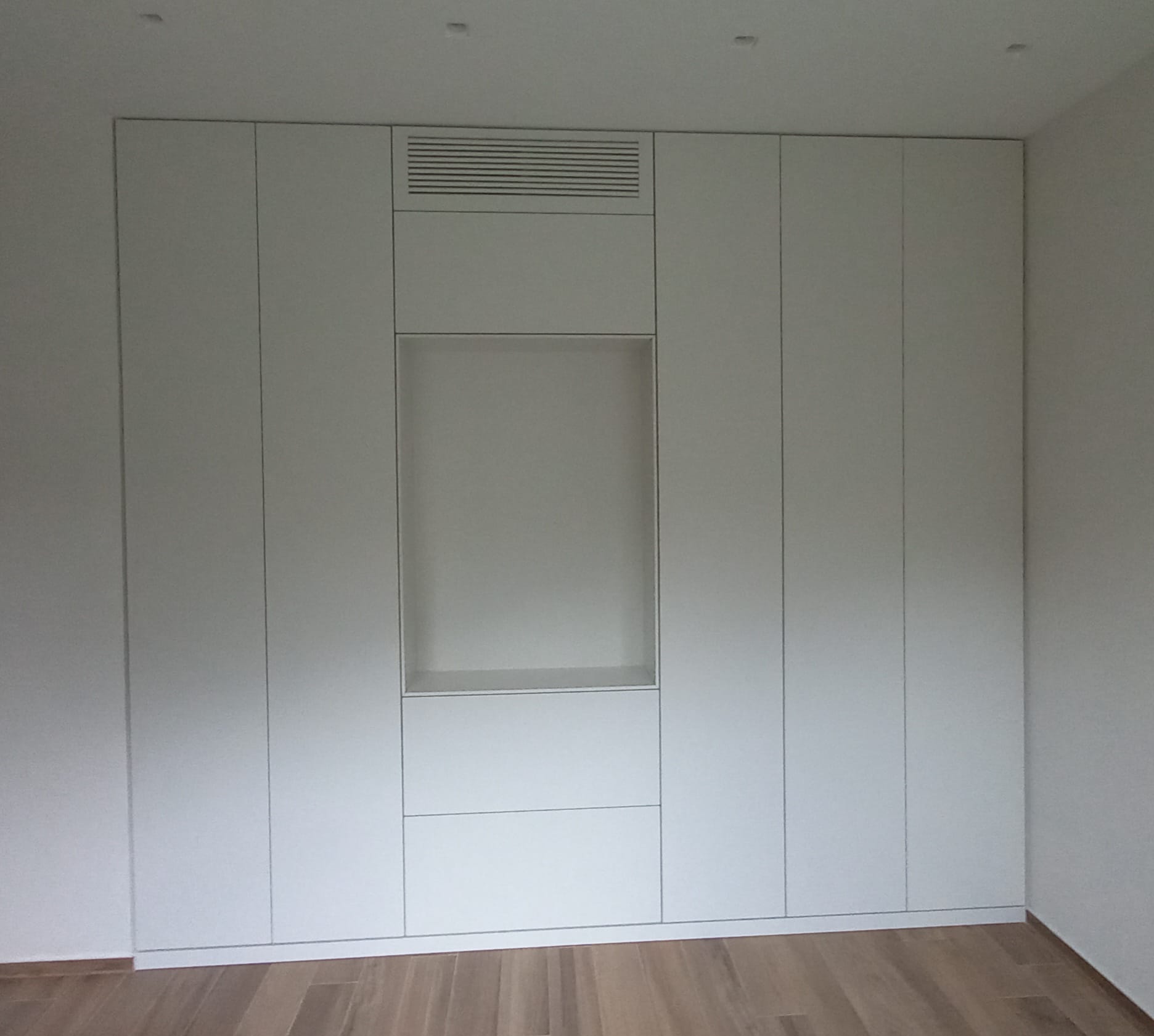 Armadio bianco a muro con nicchia rettangolare e sfiato nella parte superiore, su pavimento in legno in una stanza.