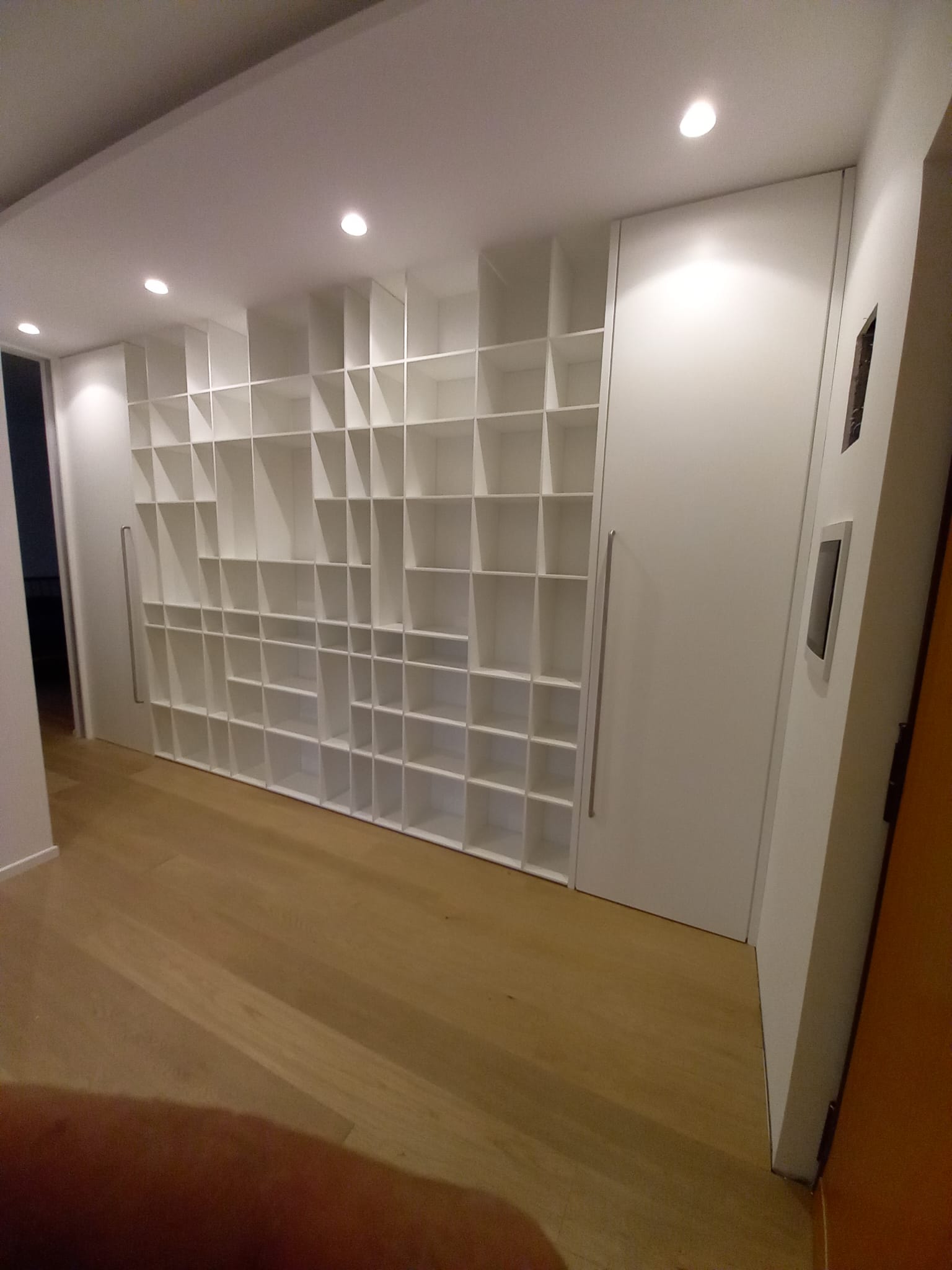 Scaffale bianco integrato con design geometrico e ante nascoste in una stanza con pavimenti in legno.