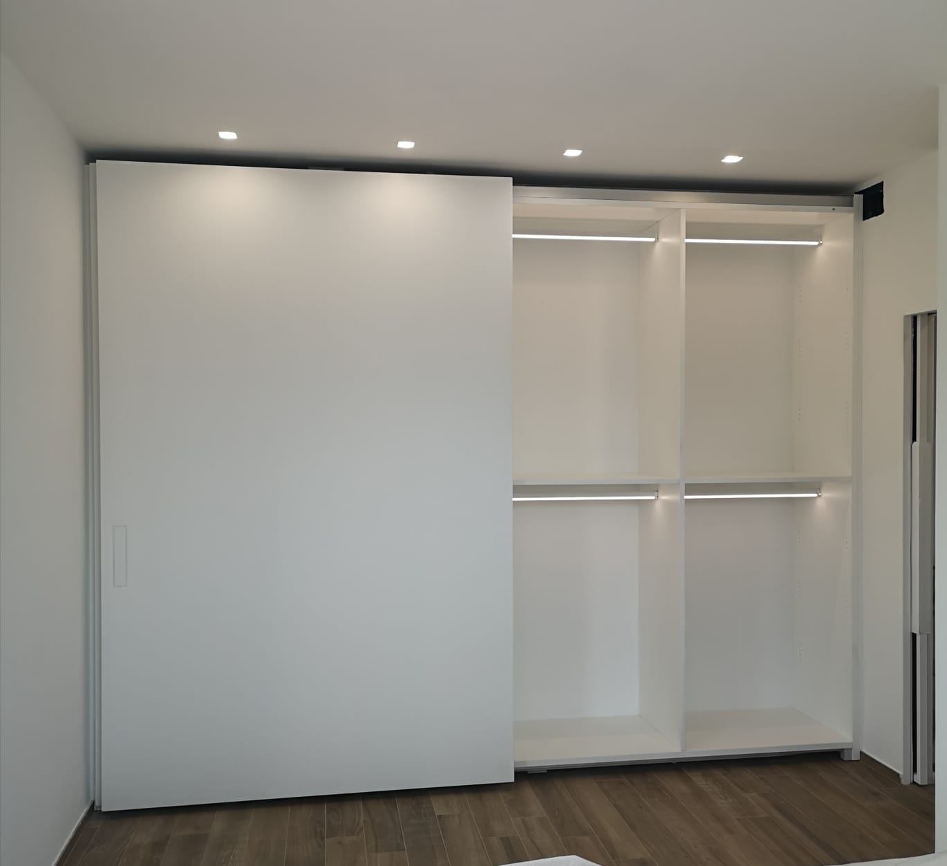 Armadio bianco con ante scorrevoli parzialmente aperto, che rivela aste appendiabiti e ripiani, illuminazione da incasso nella parte superiore.