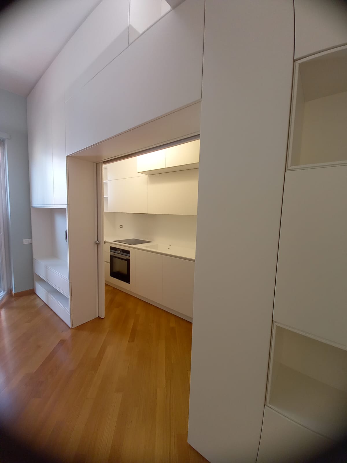 Cucina luminosa e minimalista con mobili bianchi, forno incorporato e pavimento in legno.