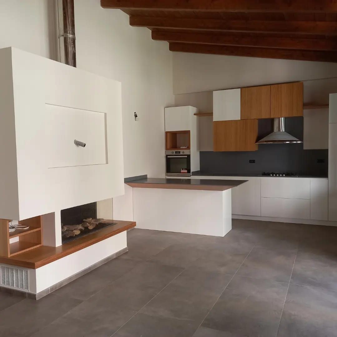 Cucina moderna con mobili bianchi, dettagli in legno, ripiani grigi e camino.