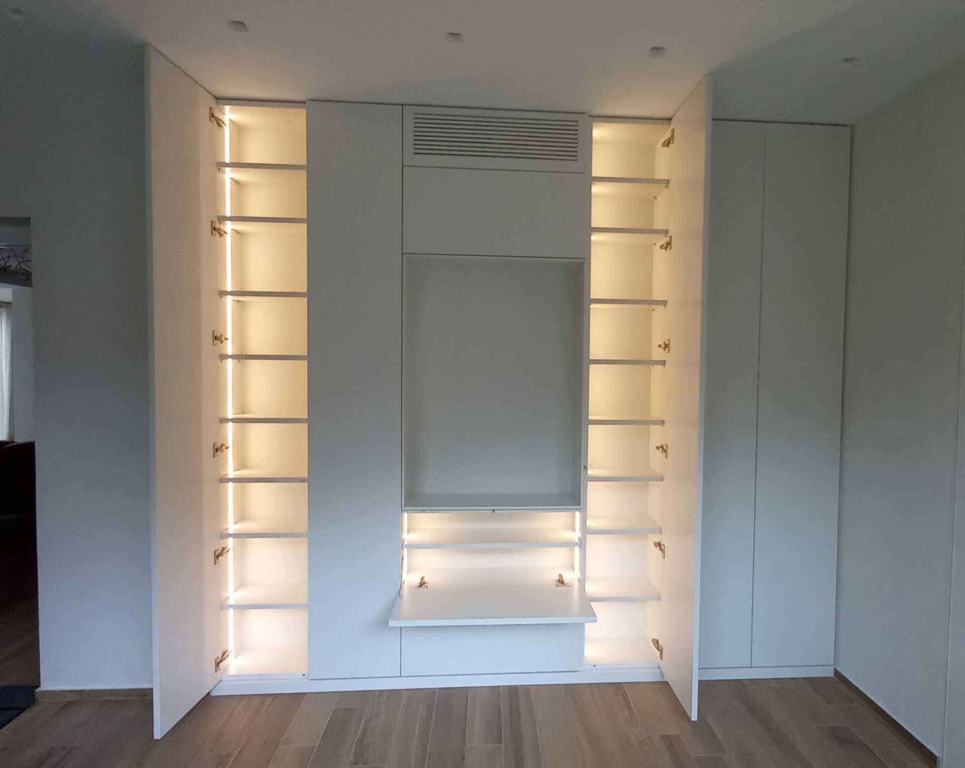 Armadio bianco a muro con ripiani illuminati, un mobile centrale e porte aperte in una stanza.