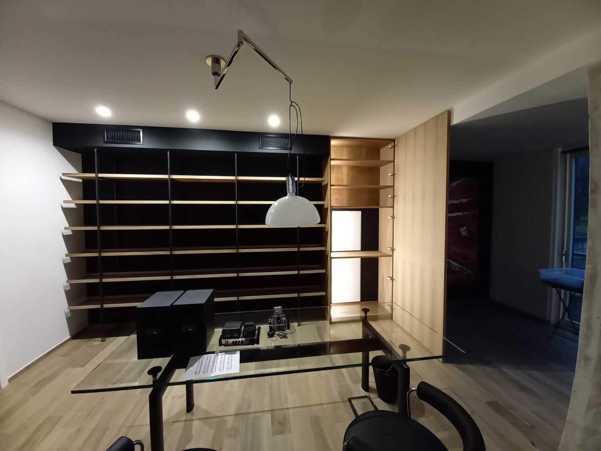 Ufficio con scaffali neri, scaffalature in legno, scrivania in vetro e lampada a sospensione.