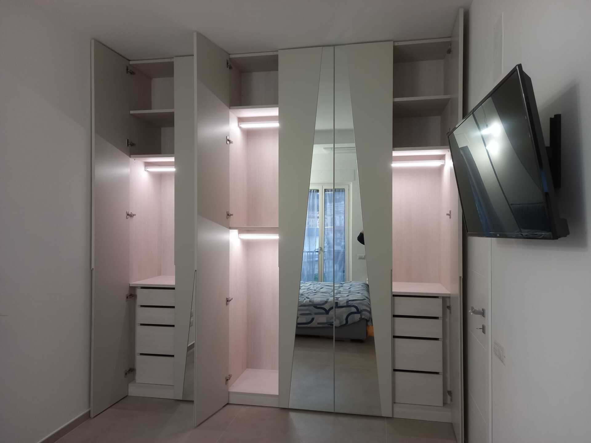 Armadio bianco con ante aperte, luci integrate, cassetti e uno specchio che riflette un letto; una TV è montata sulla parete.