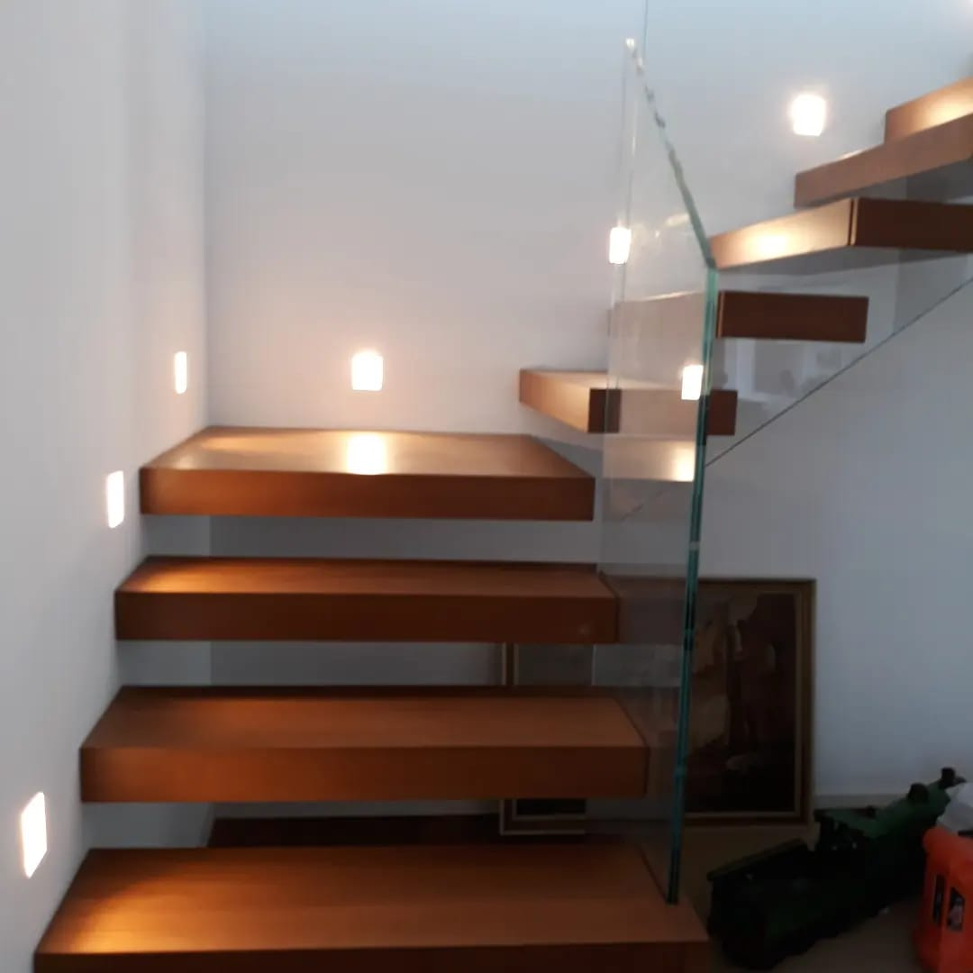 Scala in legno con luci integrate, ringhiera in vetro, che sale contro una parete bianca.