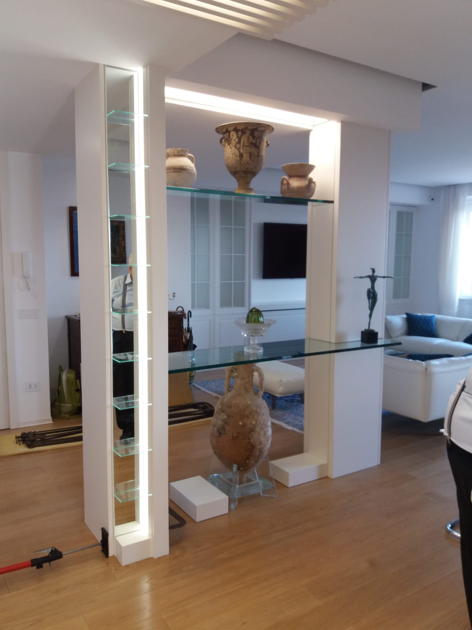 Parete divisoria bianca con ripiani in vetro su cui sono esposti vasi, sculture e illuminazione.
