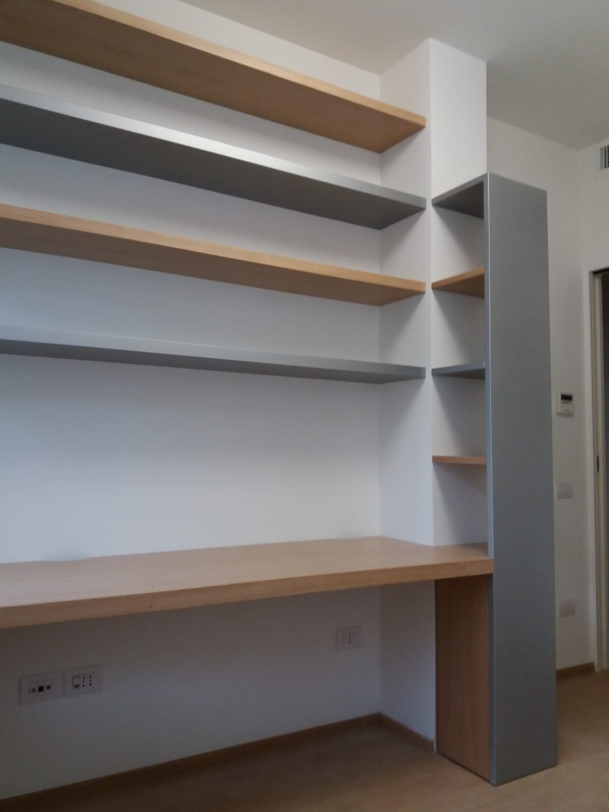 Scrivania e scaffali in legno integrati. I ripiani alternano legno grigio e naturale. Pareti bianche.