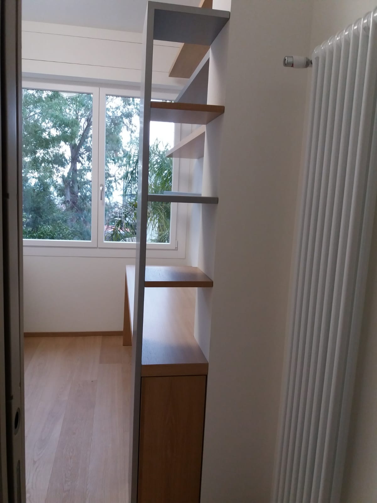 Una stretta libreria incassata divide la stanza, con ripiani in diverse tonalità di legno contro una parete bianca e una finestra che si affaccia sulla finestra.