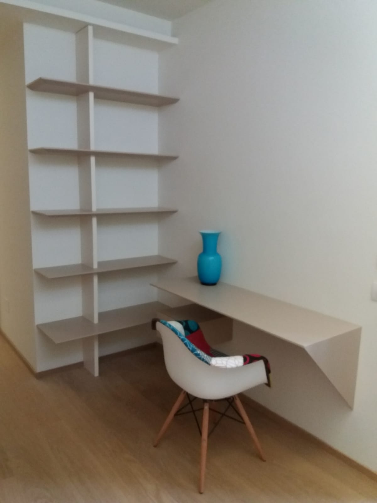 Scrivania e scaffalature integrate nell'angolo della stanza, pareti bianche, legno chiaro, vaso blu, sedia moderna.