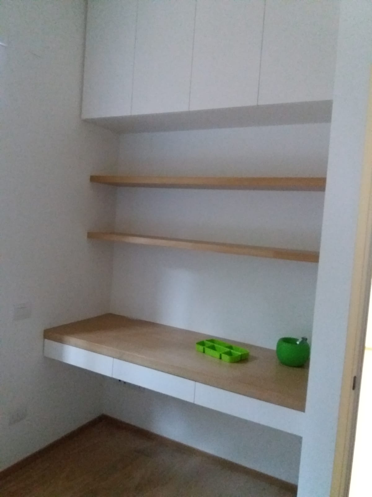 Scrivania bianca a muro con ripiani e armadietti. Superfici in legno chiaro, oggetti verdi e bianchi. Stanza vuota.