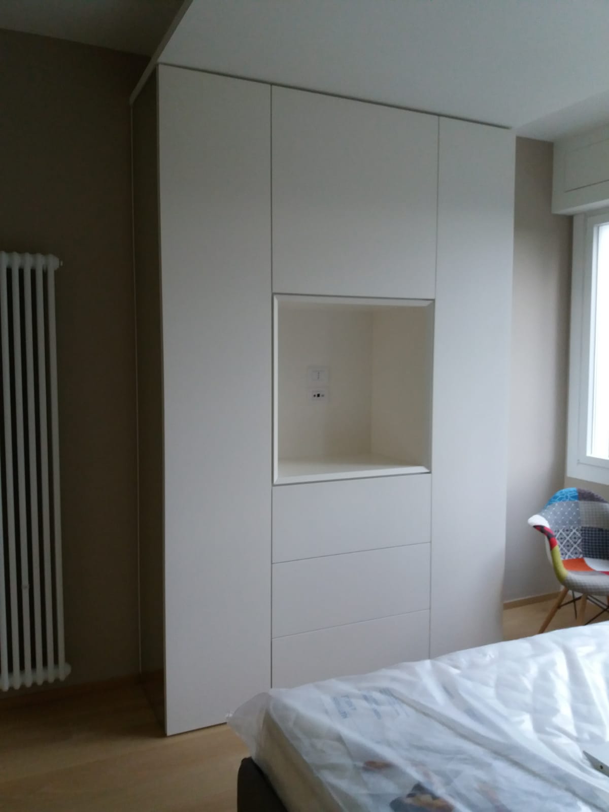 Mobile contenitore alto bianco a muro con nicchia aperta in una camera da letto. Sono visibili anche un termosifone e una finestra.
