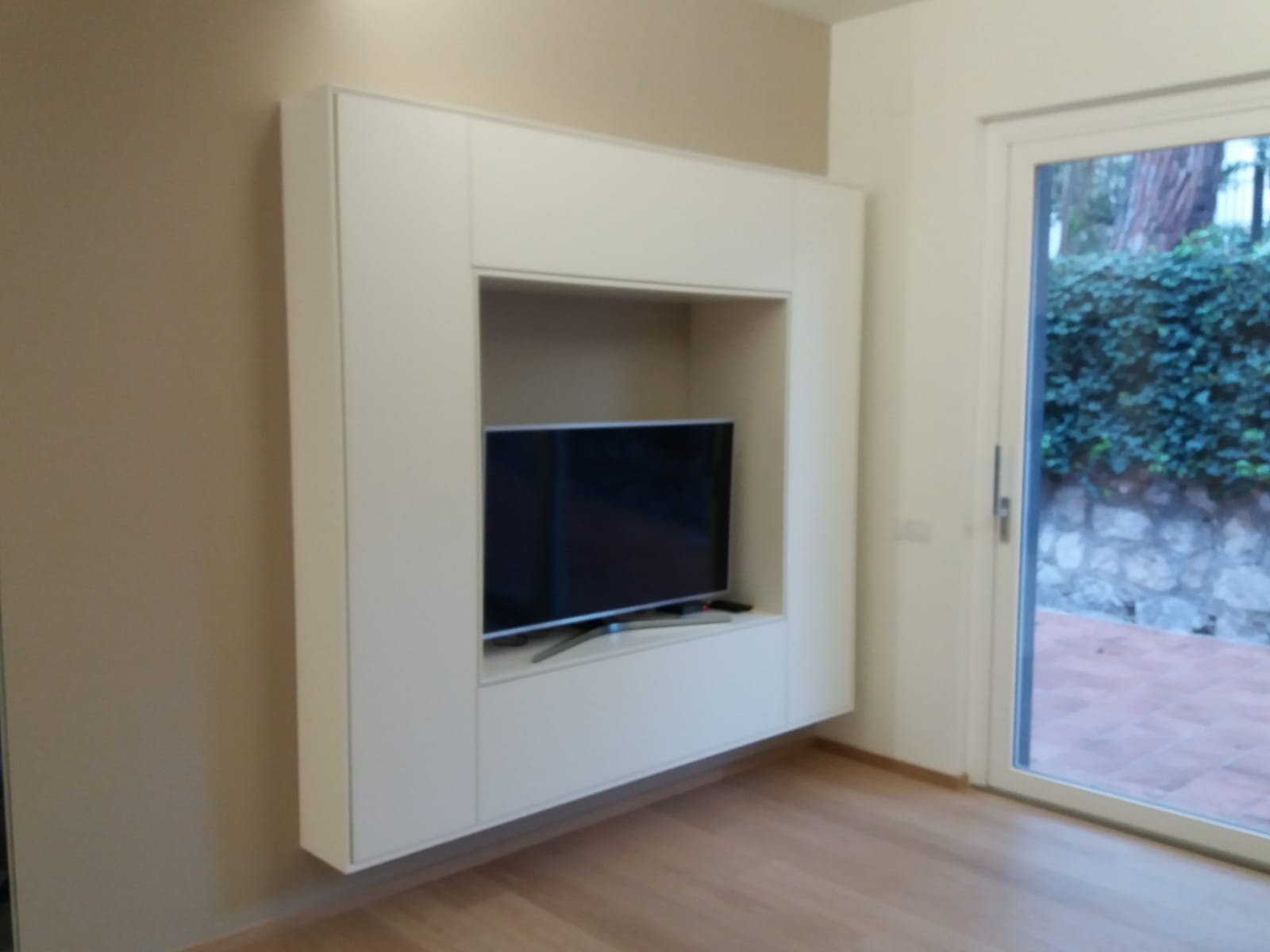 Pensile bianco con TV, montato su una parete beige. Sulla destra c'è una porta con vista sul verde.