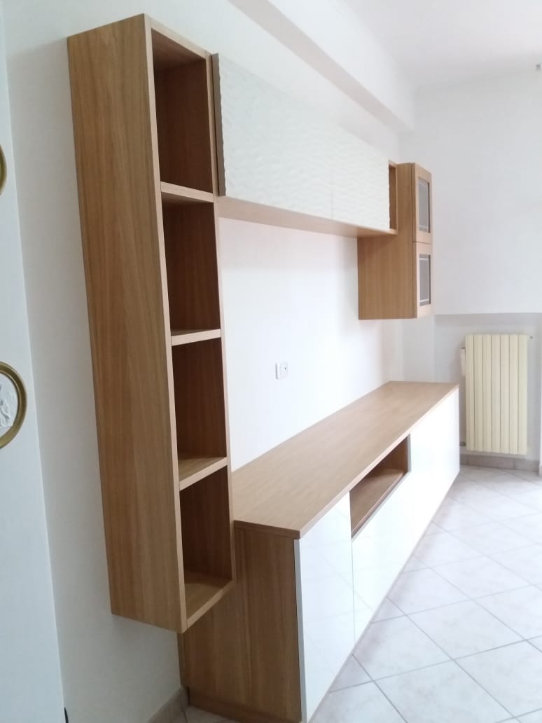 Mobile TV a parete in legno con ripiani a giorno, armadietti e una mensola sospesa. Legno chiaro contro una parete bianca.