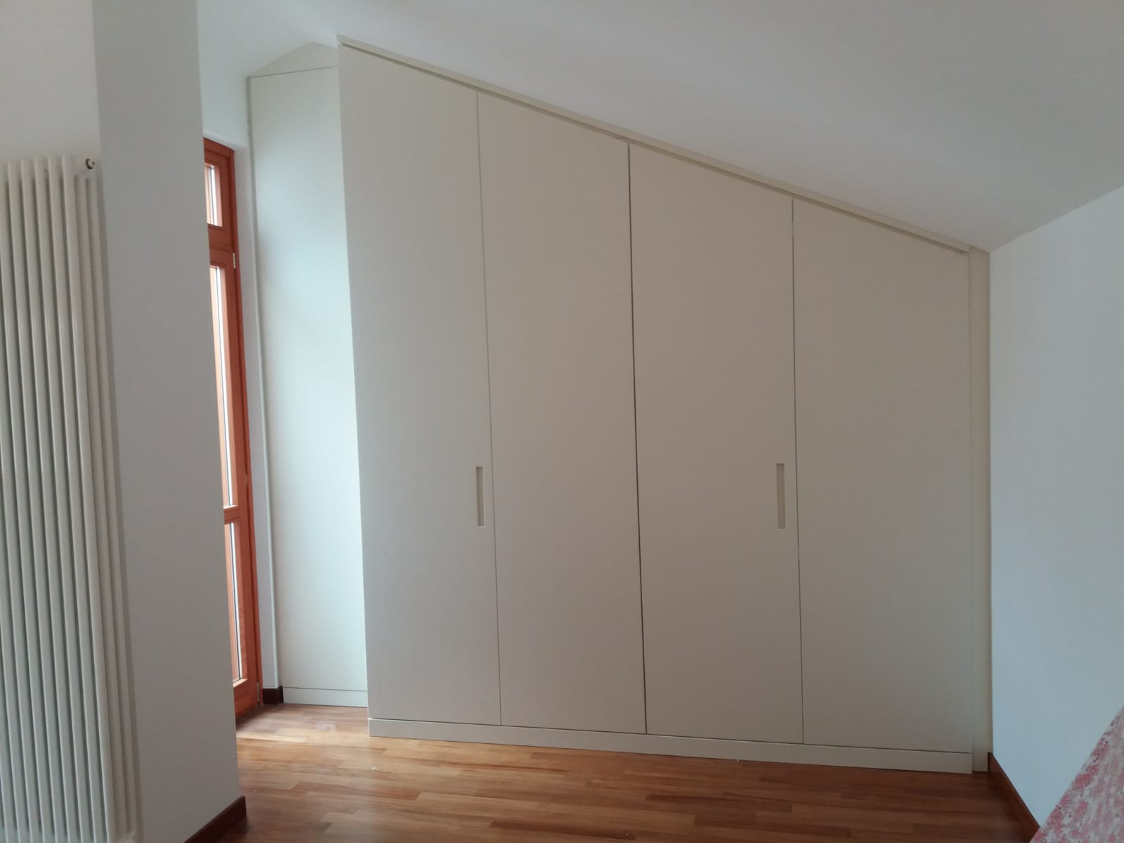 Armadio a muro color crema con piano inclinato, pavimento in legno, accanto a una finestra e un termosifone.