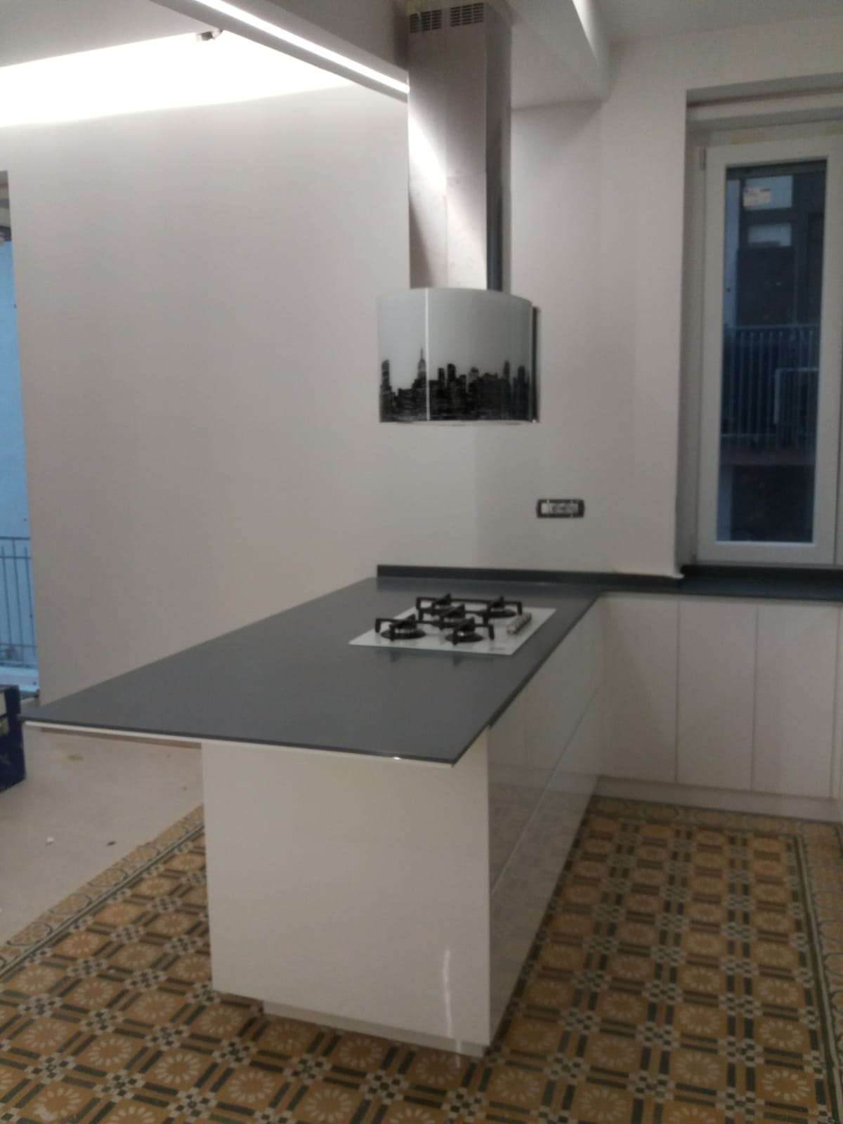 Moderna cucina bianca con isola, piano di lavoro nero, piano cottura e cappa aspirante. Finestra sulla destra.