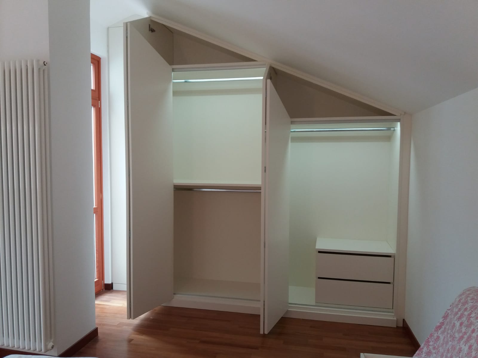 Armadio bianco a muro con soffitto spiovente in una stanza con finestra e pavimento in legno.