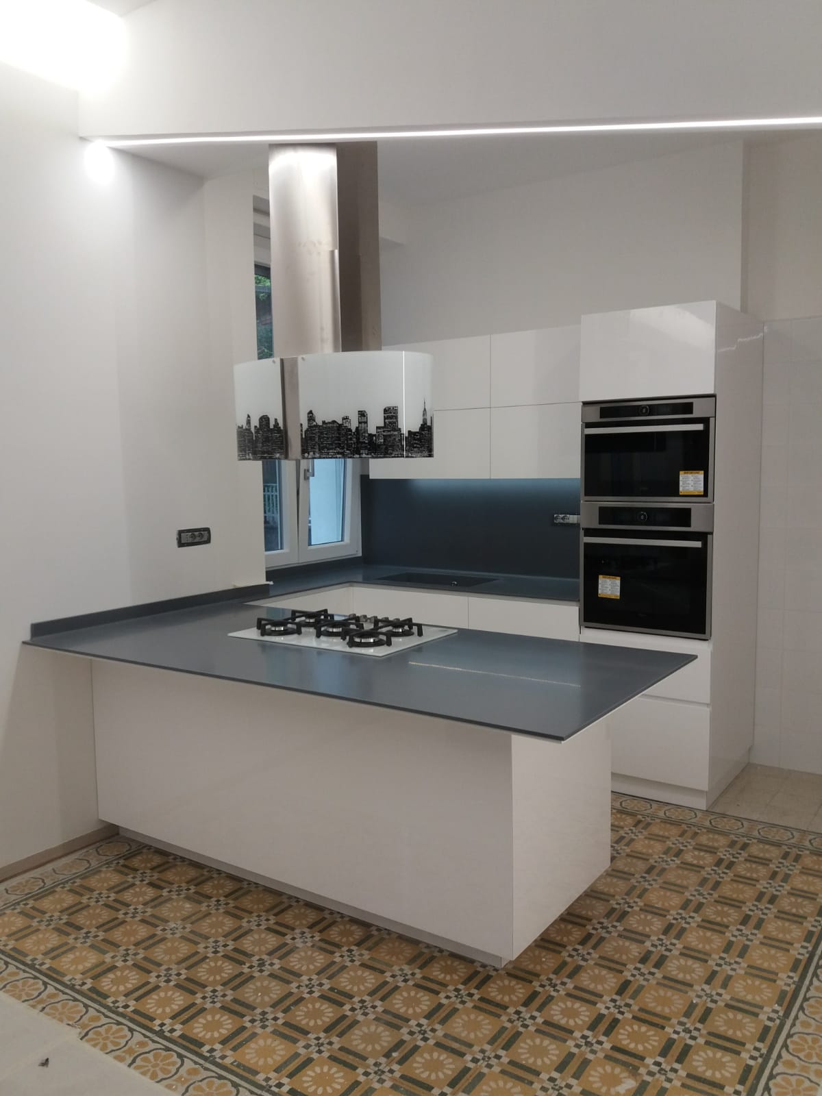 Moderna cucina bianca con isola con piano di lavoro grigio, forni integrati e pavimento in piastrelle fantasia.