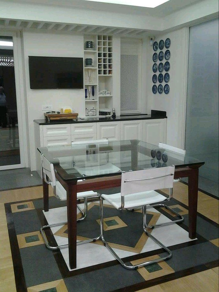 Sala da pranzo con tavolo con piano in vetro, sedie bianche, mobile con TV, piatti decorativi alla parete e tappeto a fantasia.