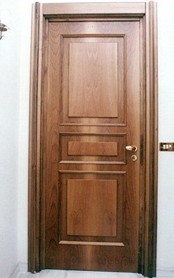 Porta in legno con tre pannelli incassati, ferramenta in ottone, inserita in un telaio in legno contro una parete bianca.
