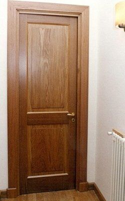 Porta in legno con pannelli, maniglia dorata, situata in un corridoio chiaro con un termosifone sulla destra.