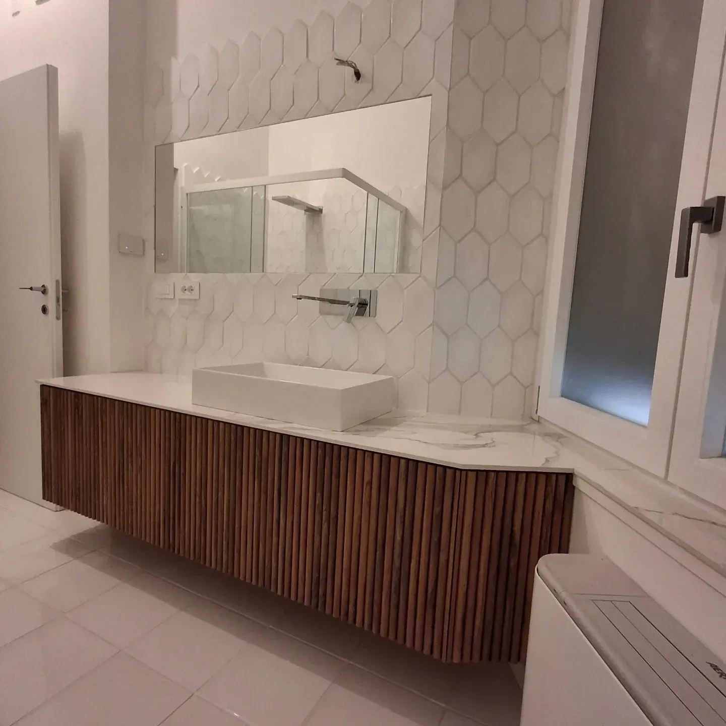 Bagno moderno con mobile in legno, lavandino bianco, parete con piastrelle esagonali, specchio e finestra.