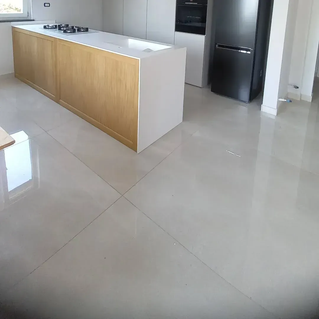 Cucina con isola in legno chiaro, piano di lavoro bianco e pavimento piastrellato grigio; frigorifero a vista.