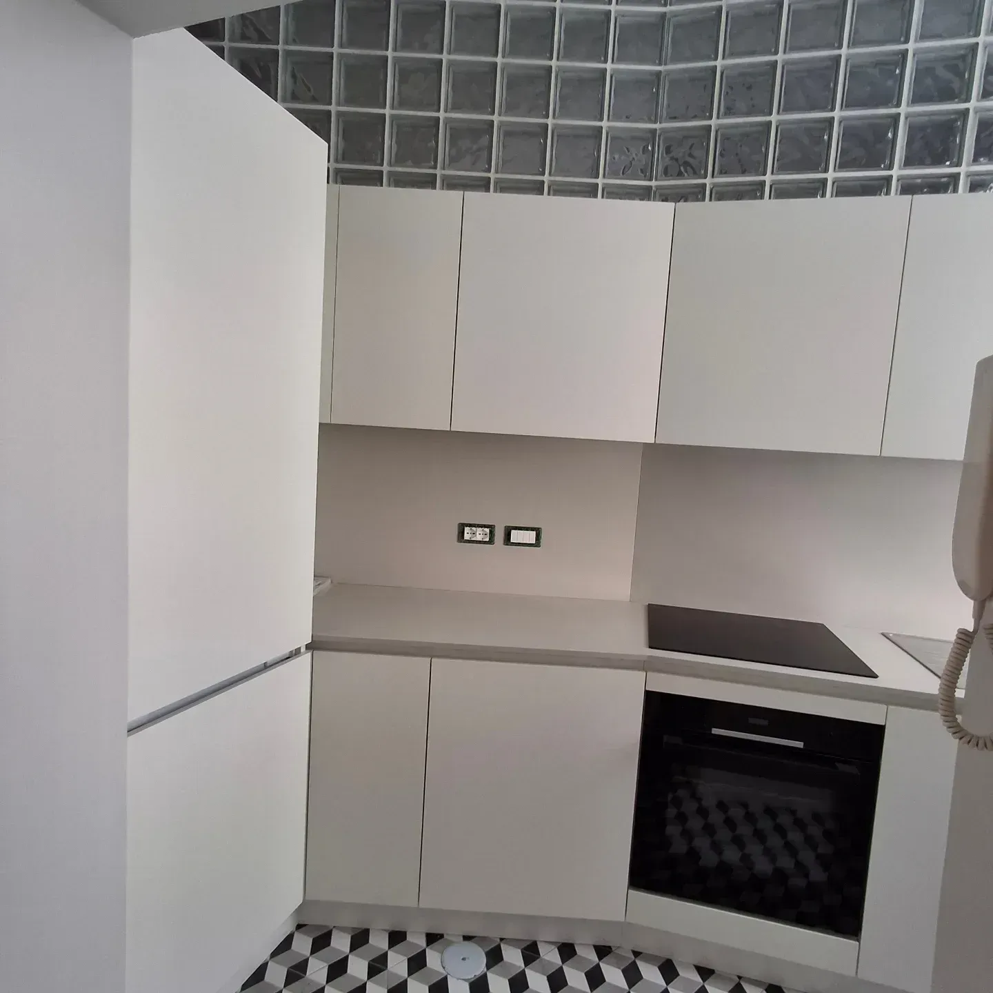 Moderna cucina bianca con forno nero, bancone e paraschizzi grigi, pavimento a scacchi e dettagli in piastrelle di vetro.