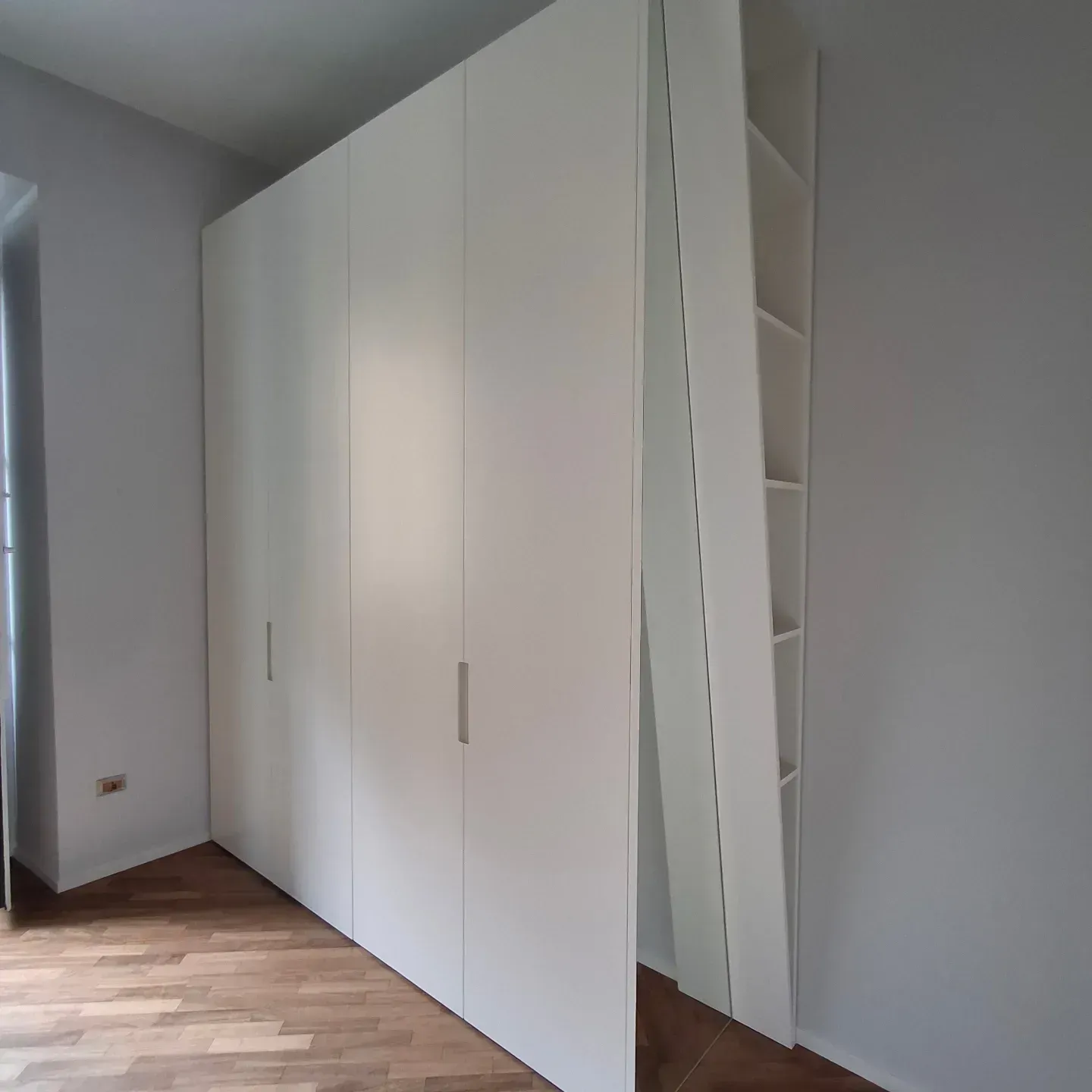 Armadio a muro bianco con sei ante. Un'anta è aperta e rivela dei ripiani. Stanza con pavimenti in legno.