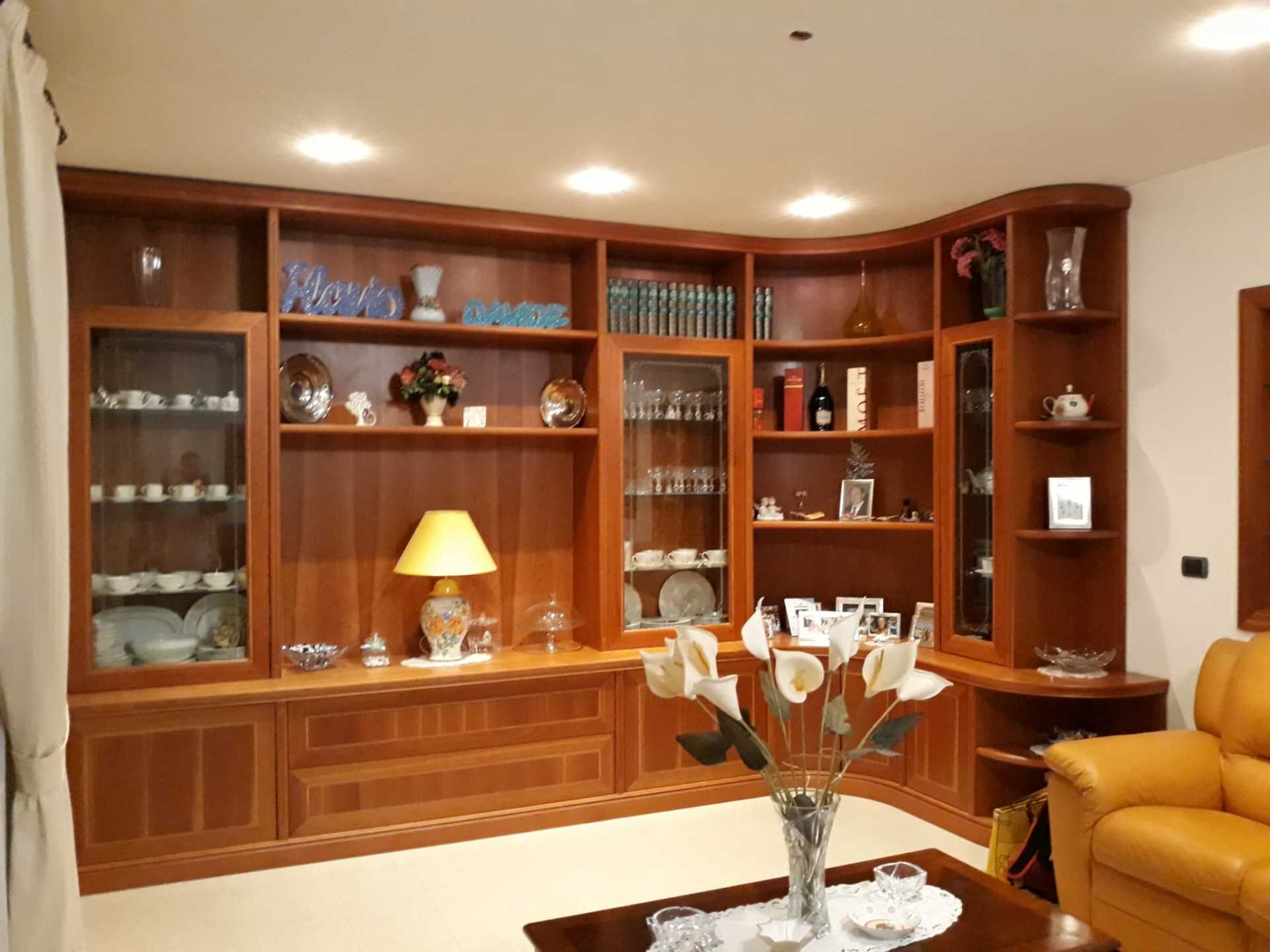 Scaffale in legno integrato, curvo, pieno di oggetti. Include armadietti, ante in vetro, libri e oggetti decorativi.