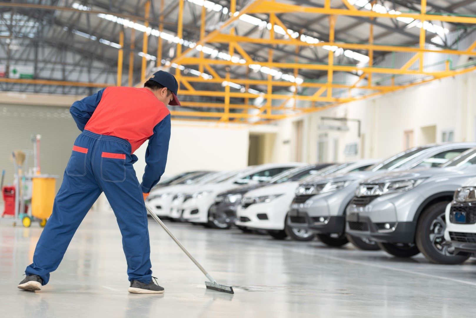 A person in blue overalls and red vest sweeping a floor in a car dealership.