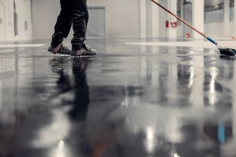 Person applying sealant to a shiny, dark floor in a large, industrial space.