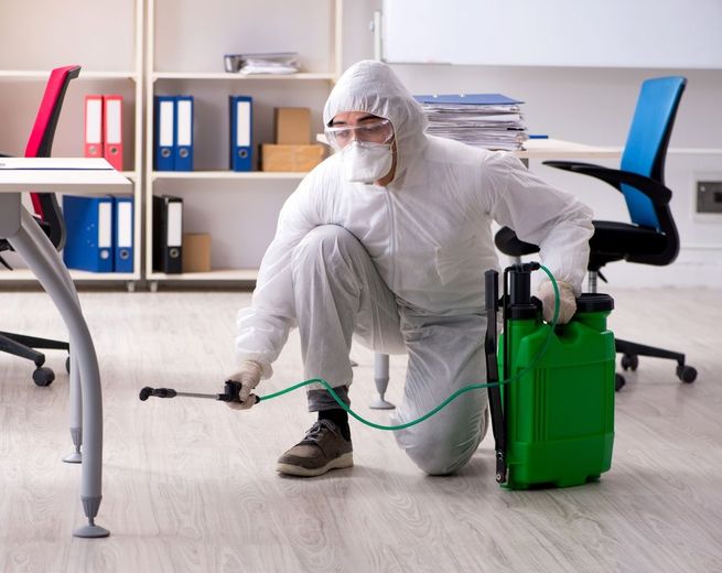 A man in a protective suit is disinfecting an office with a sprayer.