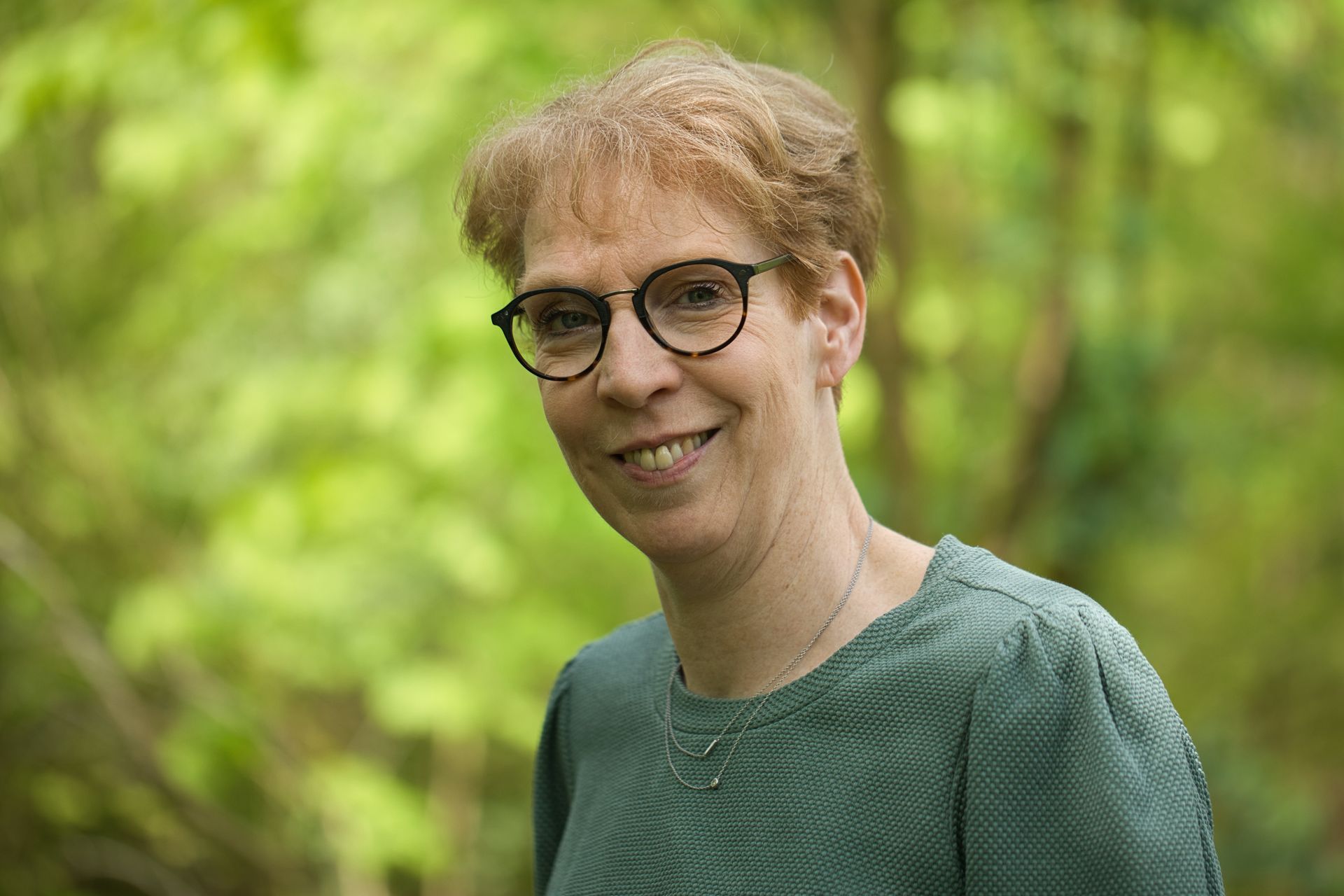 Vrouw met bril lacht buiten, groene trui, bomen wazig op de achtergrond.