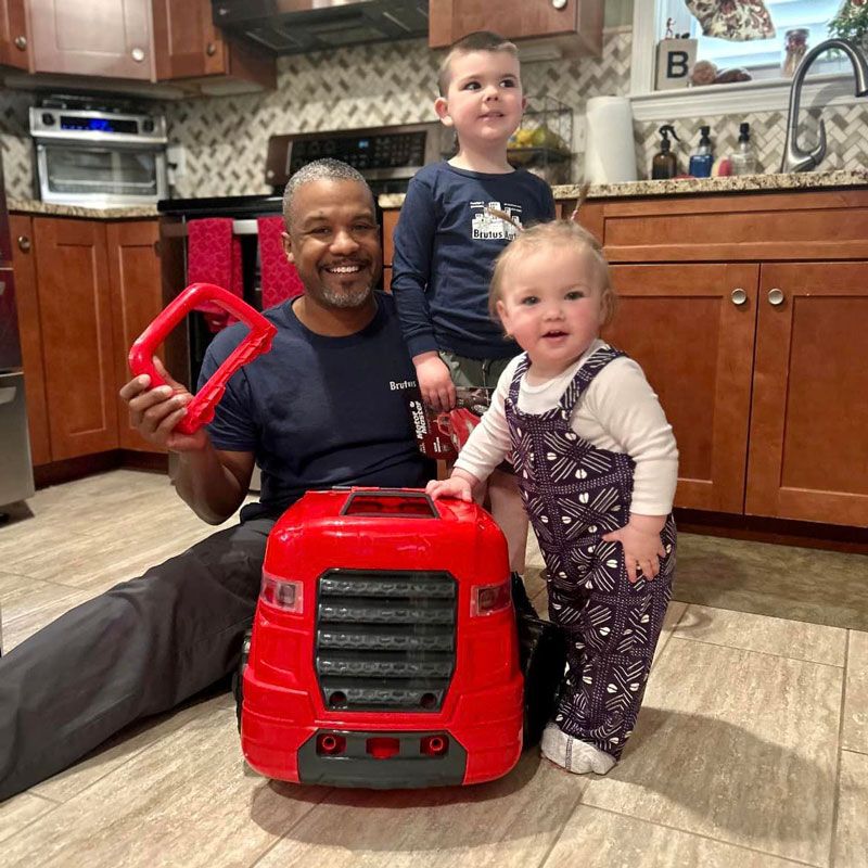 A man and two children are playing with a toy tractor in a kitchen.