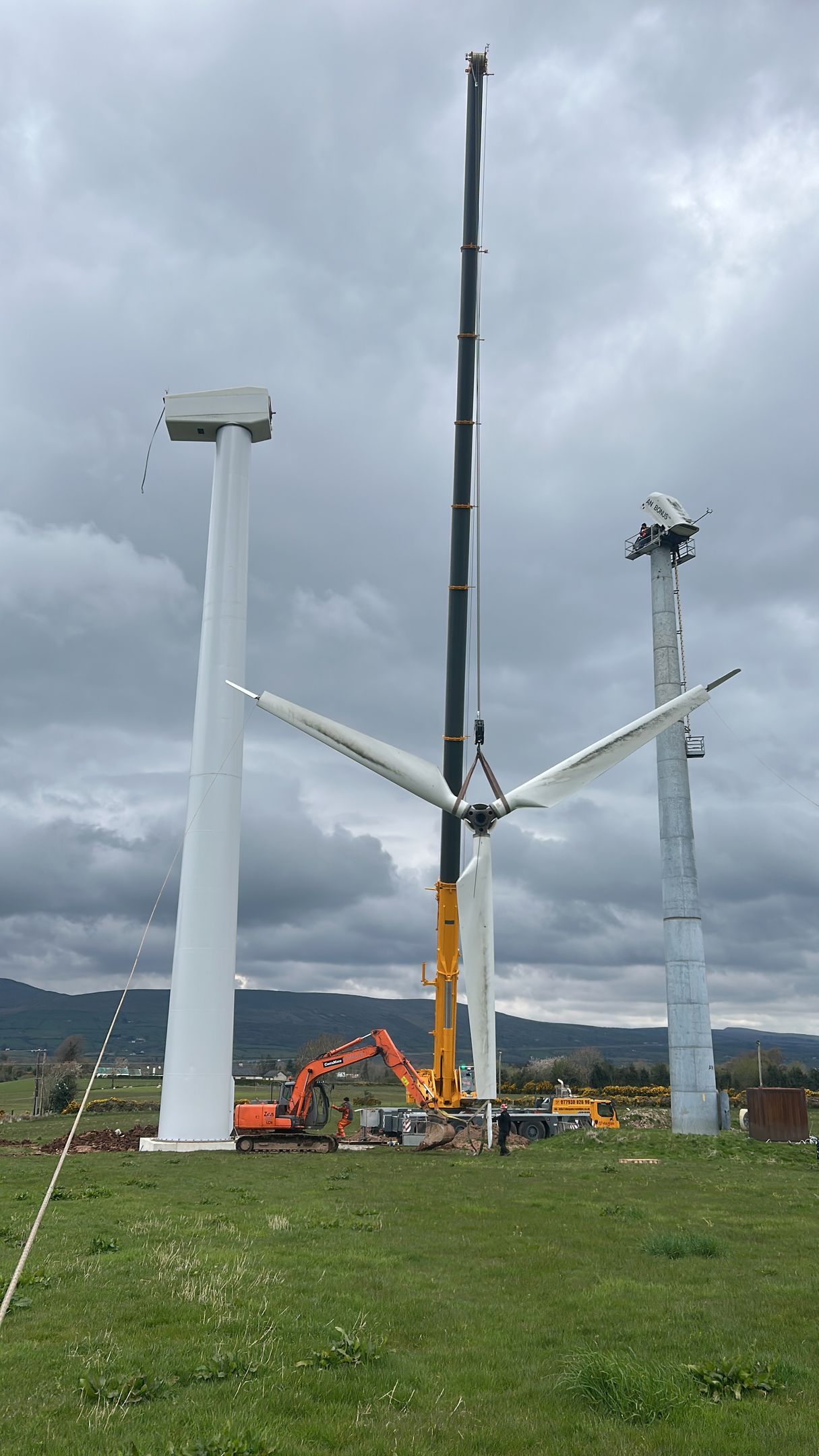 Wind turbine under construction with tall crane and service trucks in a grassy field under cloudy sky