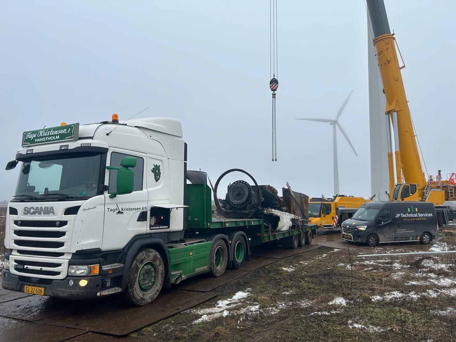 Hijskraanapparatuur naast een witte vrachtwagen en een bestelbus op een modderig bouwterrein voor een windmolenpark.