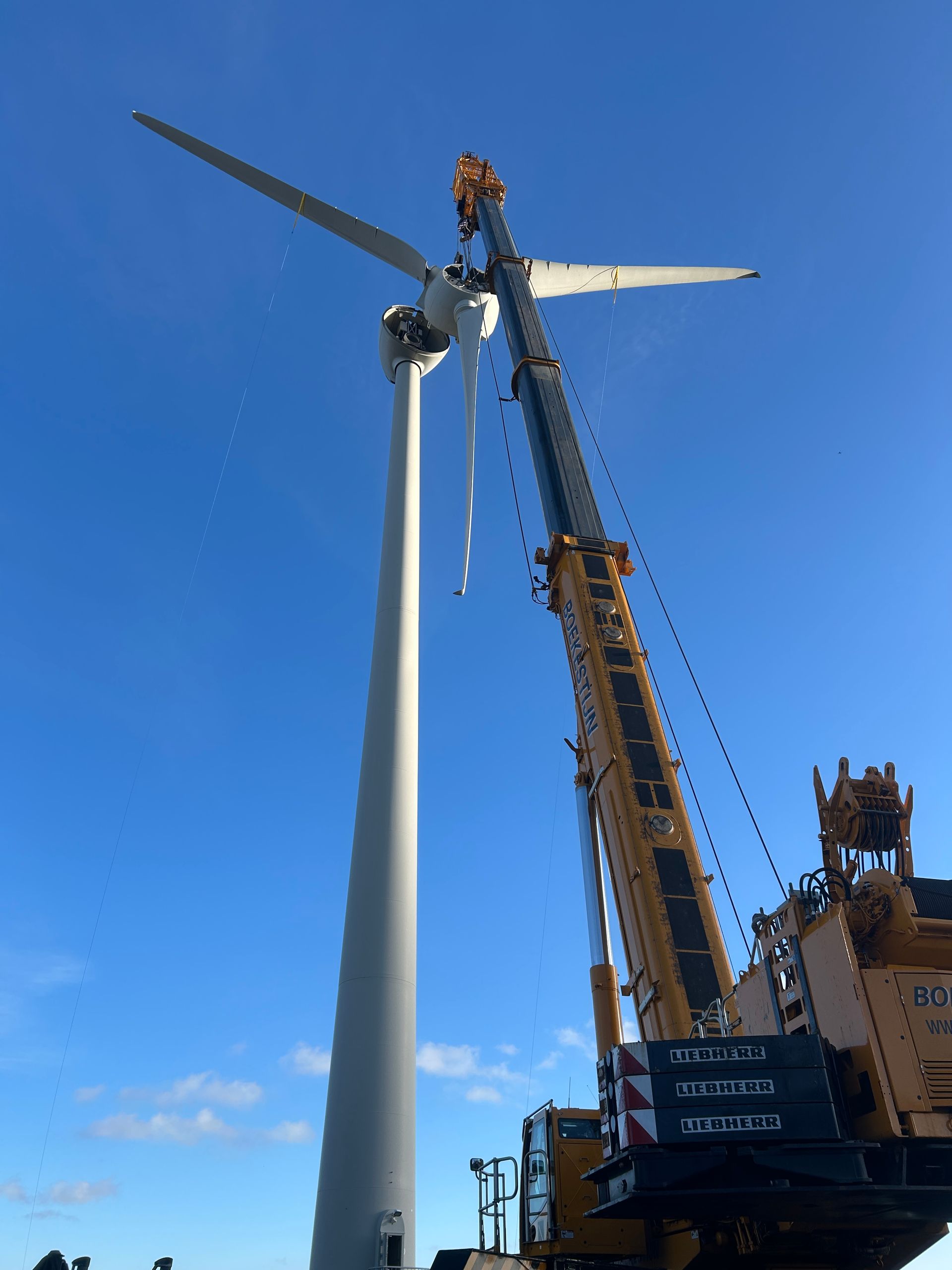 Crane installing a wind turbine blade on a white tower against a clear blue sky
