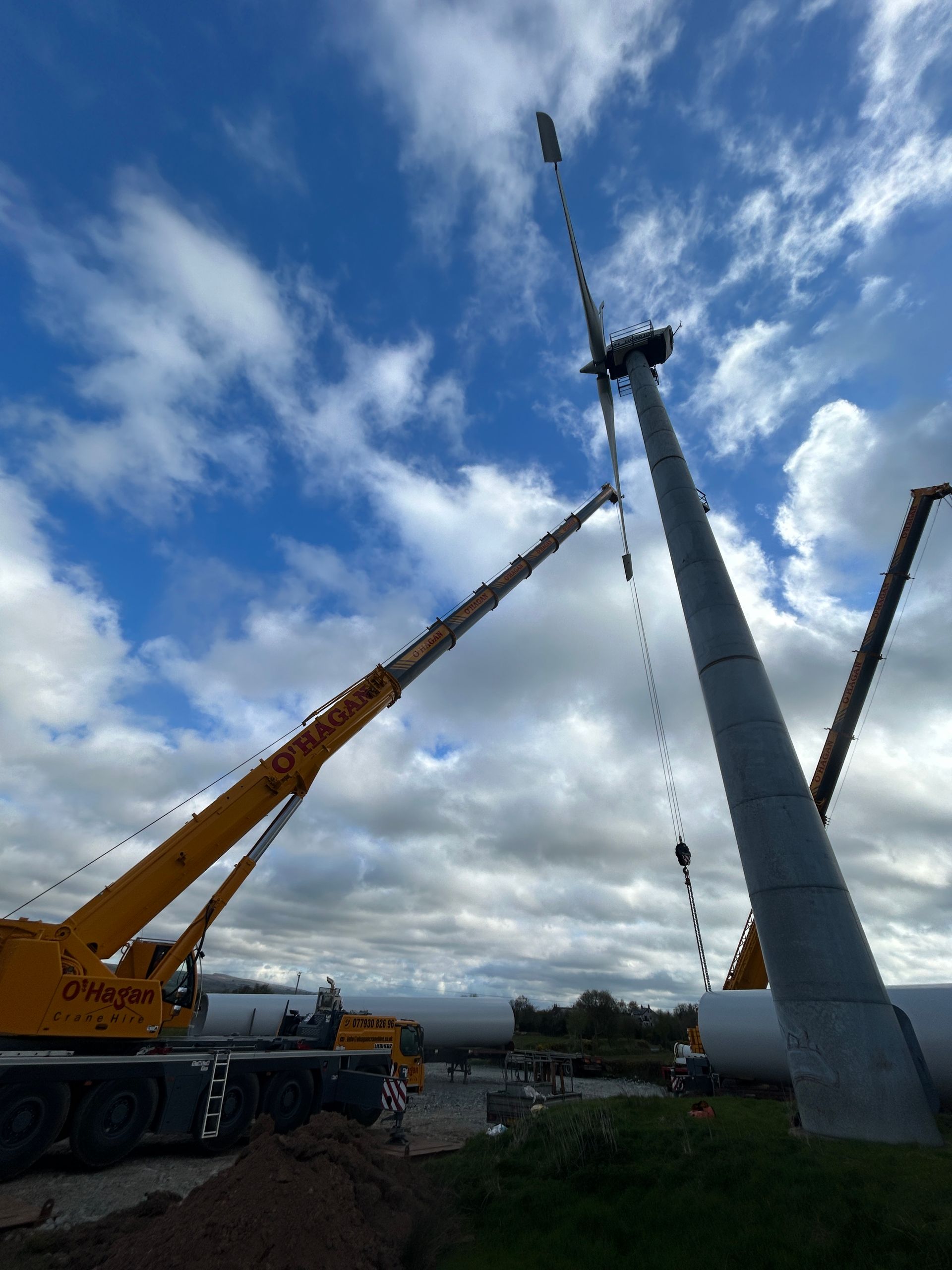Crane lifting a tall gray mast into place under a cloudy sky, with another crane nearby.