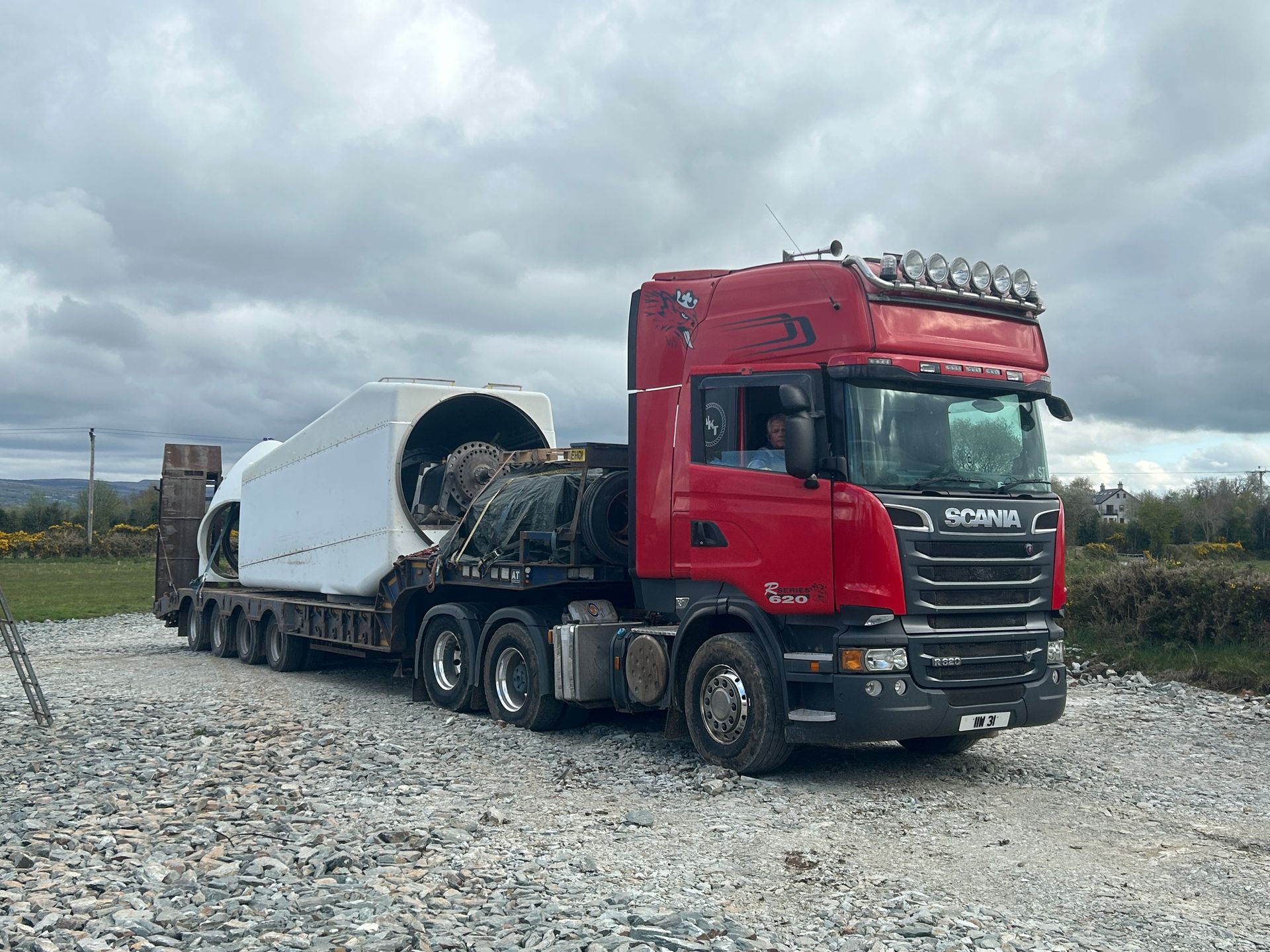 Red semi truck hauling a large white industrial component on a gravel road under cloudy skies