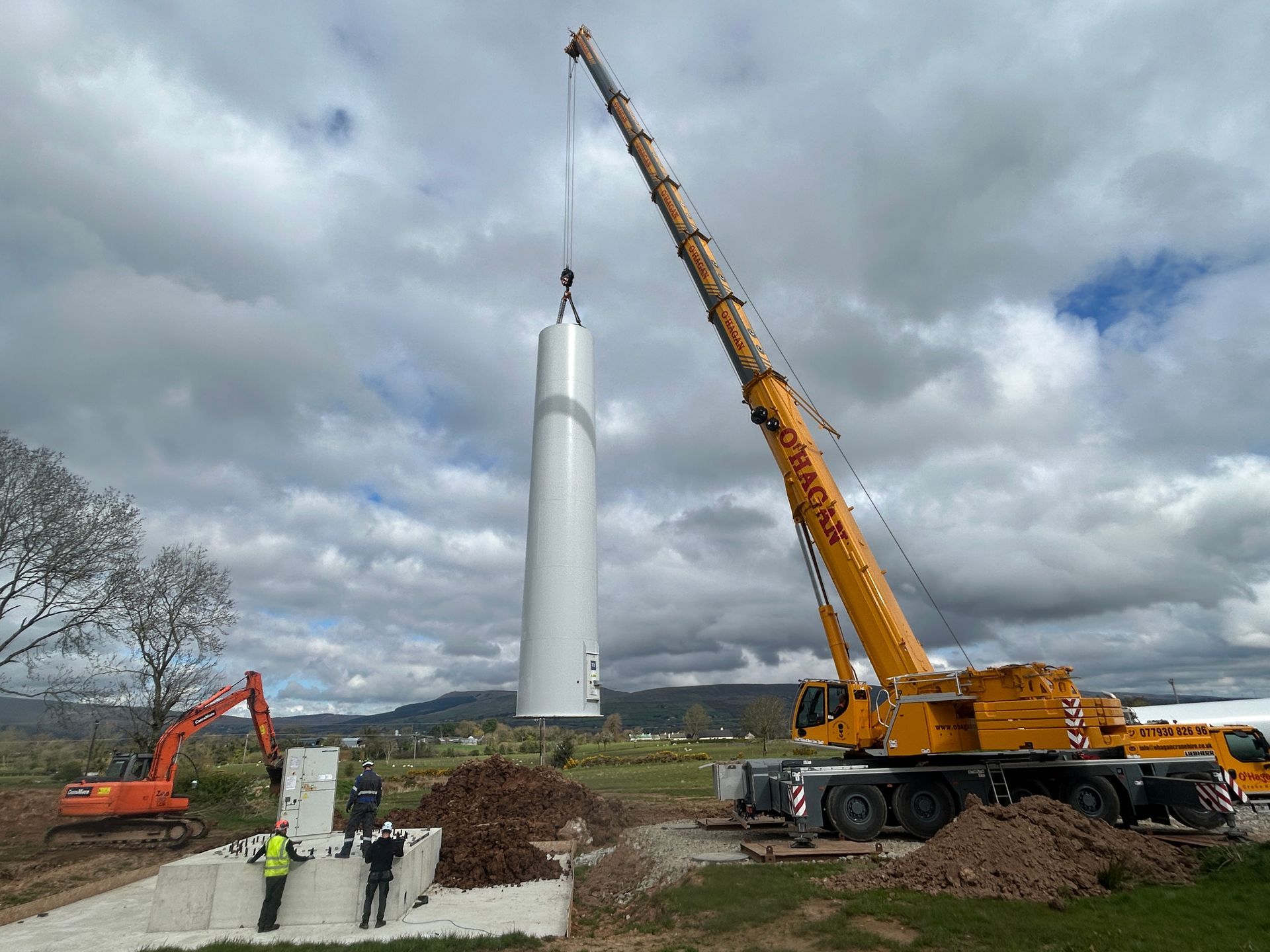 Crane lifting a tall white wind turbine tower section at a construction site under cloudy skies