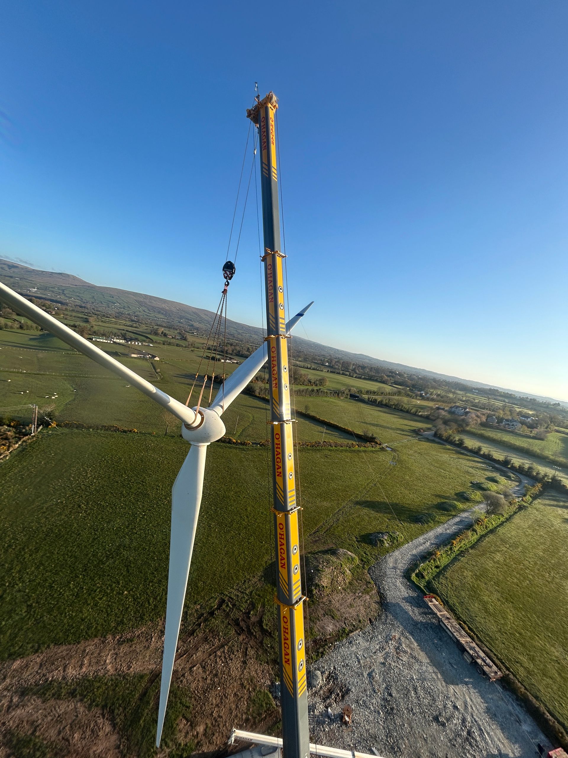 Wind turbine beside a yellow tower on a grassy plain, with a winding road and blue sky.