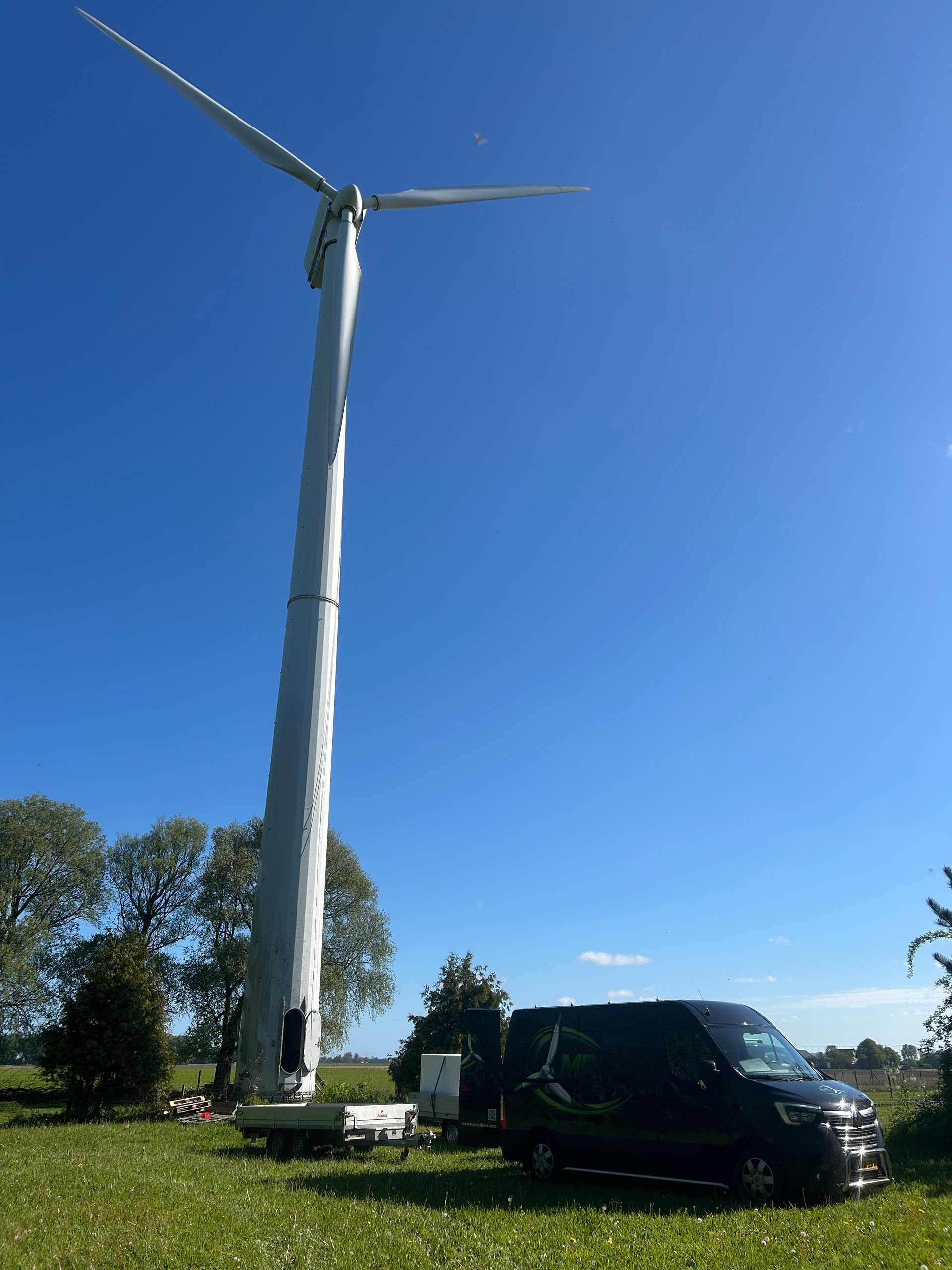 Tall wind turbine beside a black van on a sunny grassy field under a clear blue sky. Dismantling Micon 700-225  Fryslân The Netherlands