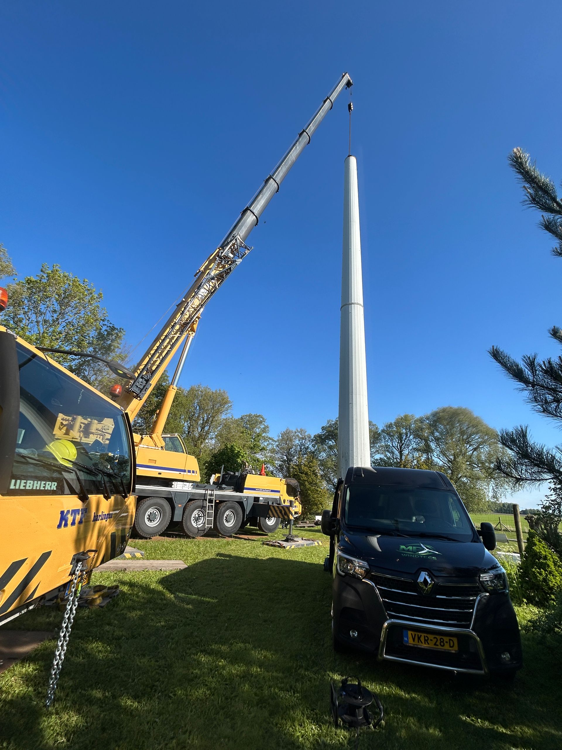 Crane lifting a tall white wind turbine blade beside a black van under a clear blue sky