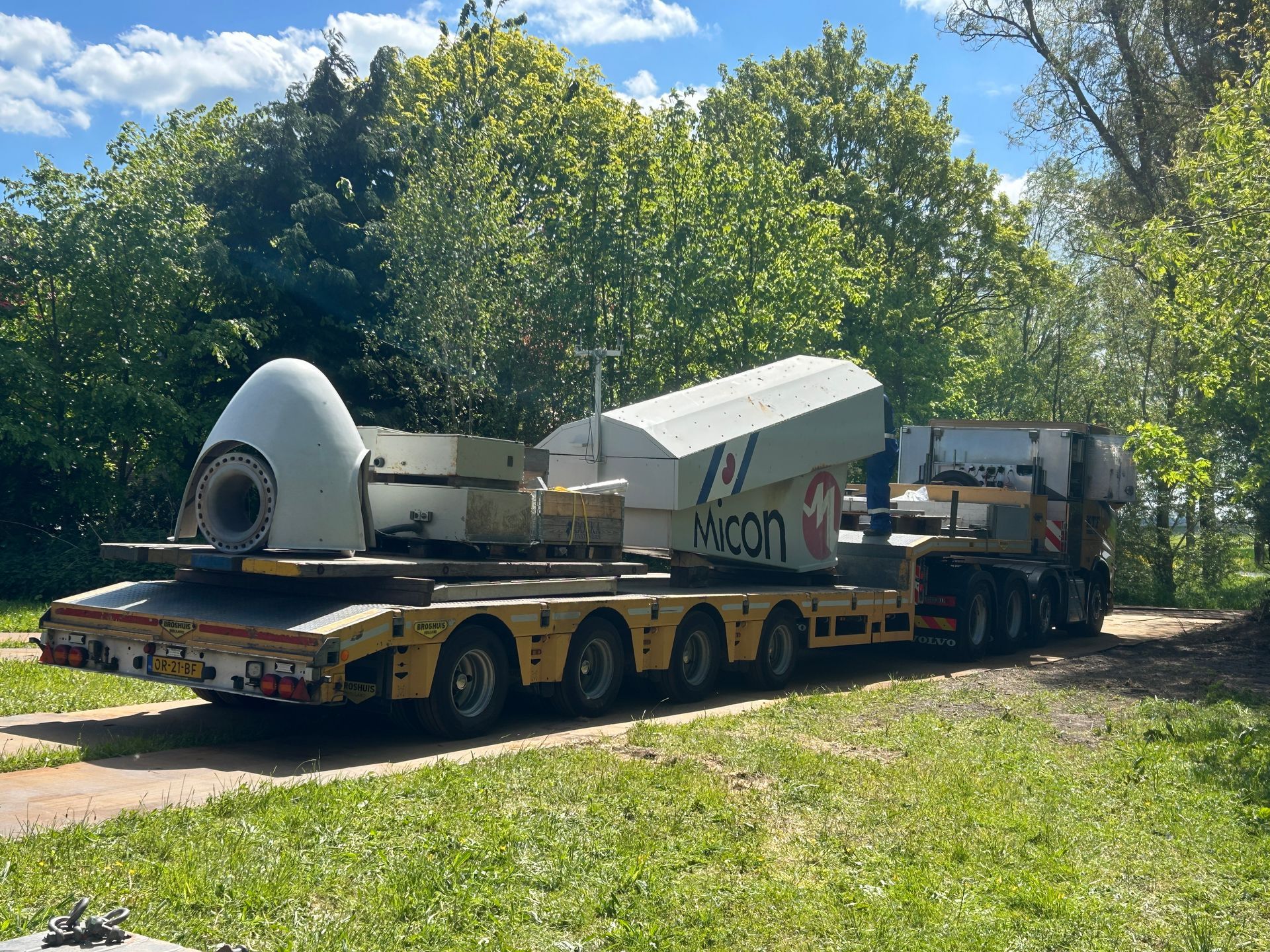 Oversized industrial machine on a flatbed trailer being hauled down a wooded dirt road