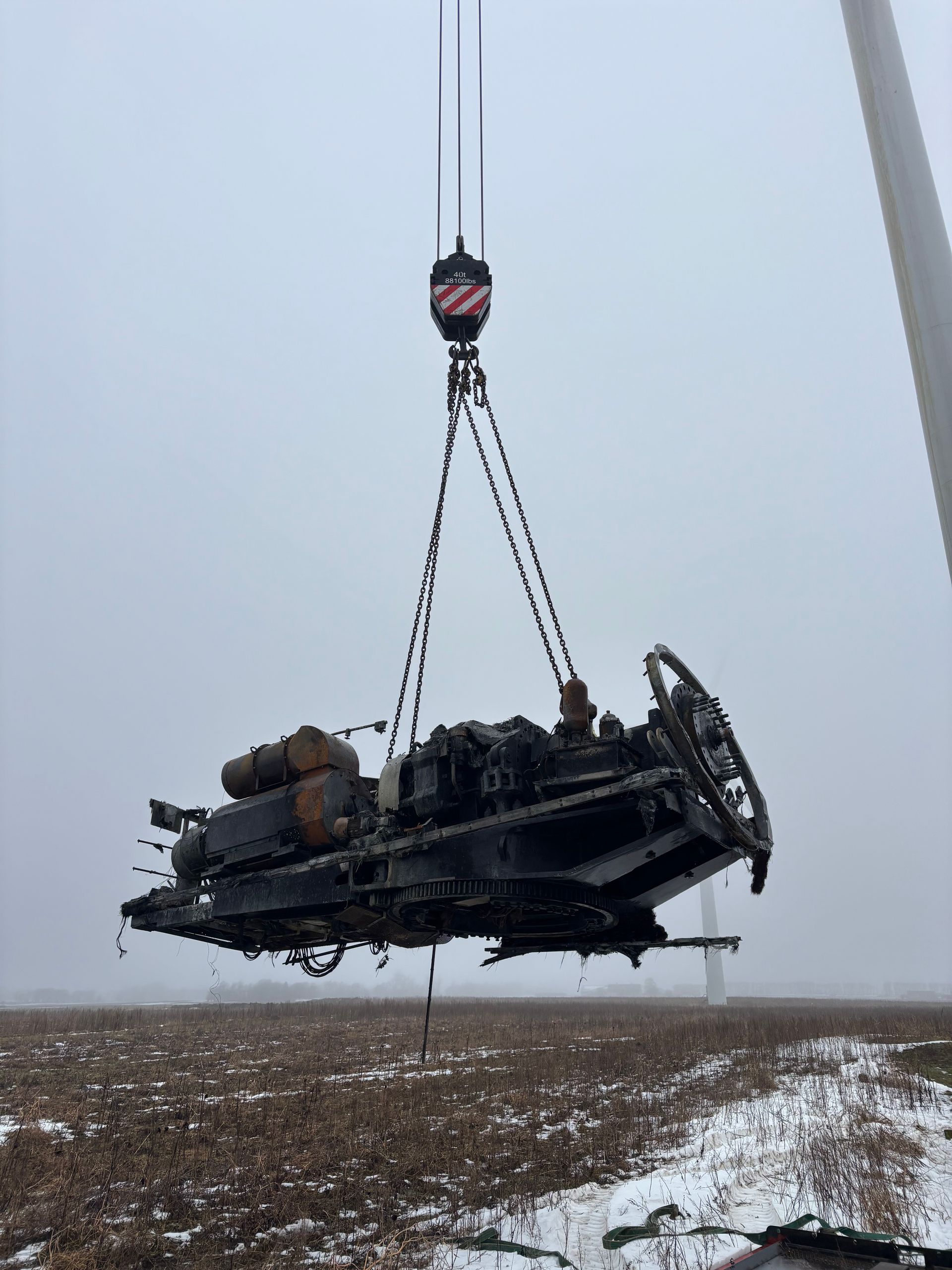 Een kraan tilt een verongelukt zwart voertuig over een besneeuwd veld in de mist.