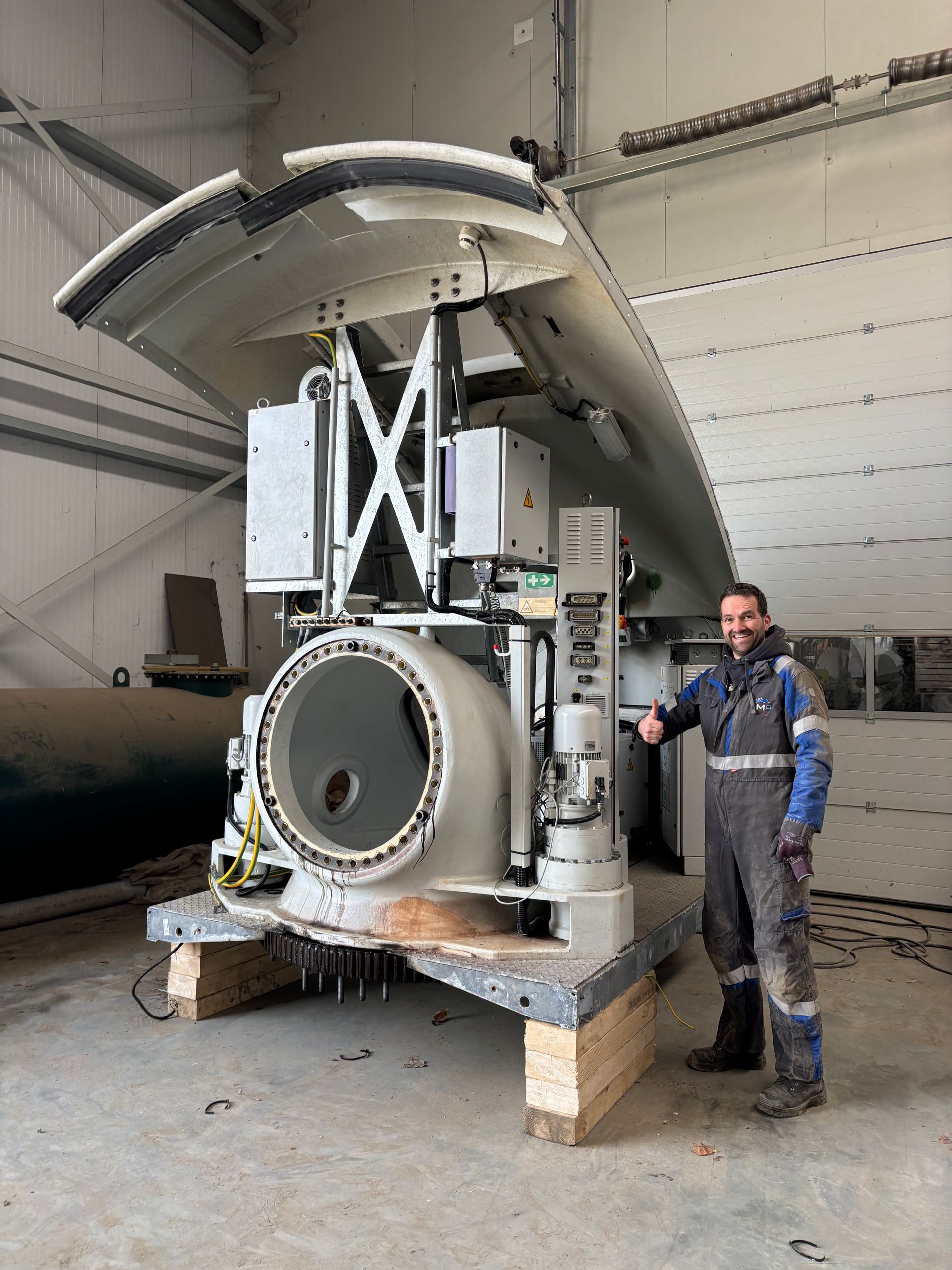 Een man staat naast een grote industriële machine met een open, gebogen bovenkant in een magazijnachtige ruimte.