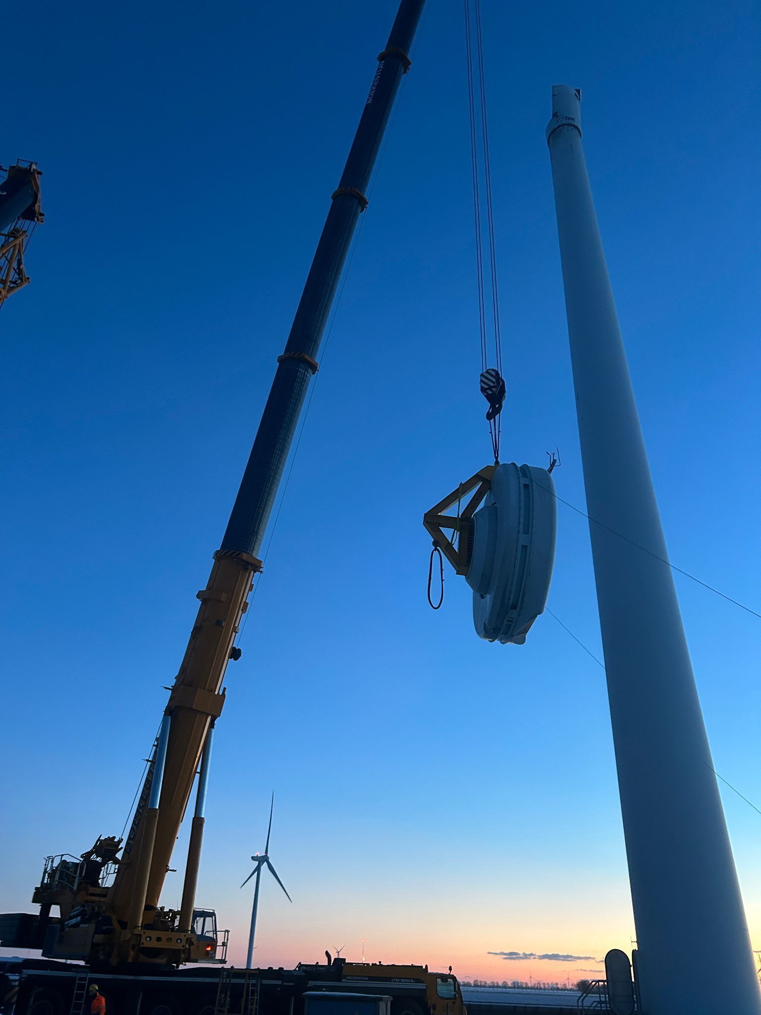 Crane lifting a large turbine blade against a blue dusk sky, with a wind turbine in the distance.