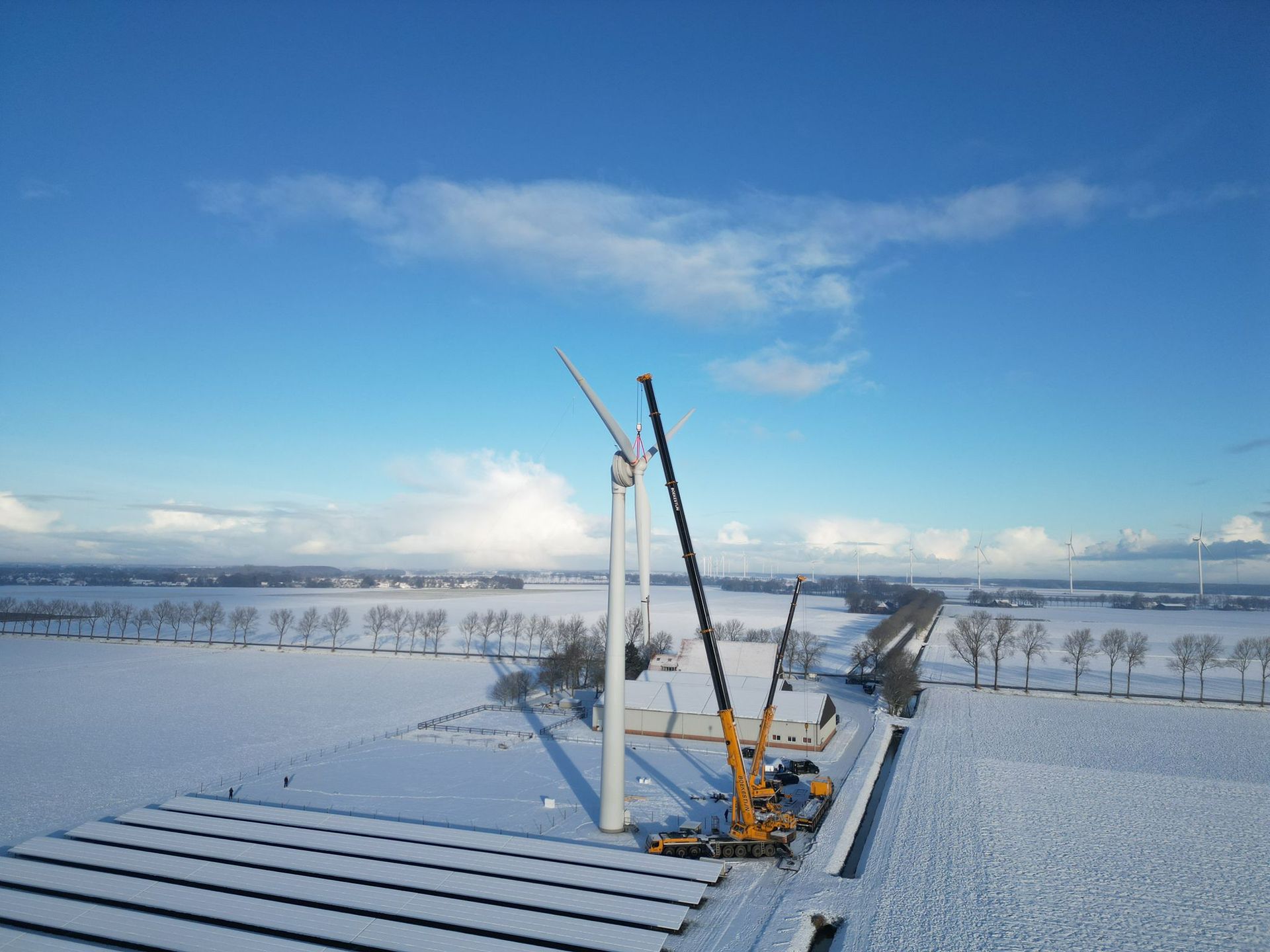 Snowy field with wind turbine components and a crane under a blue sky
