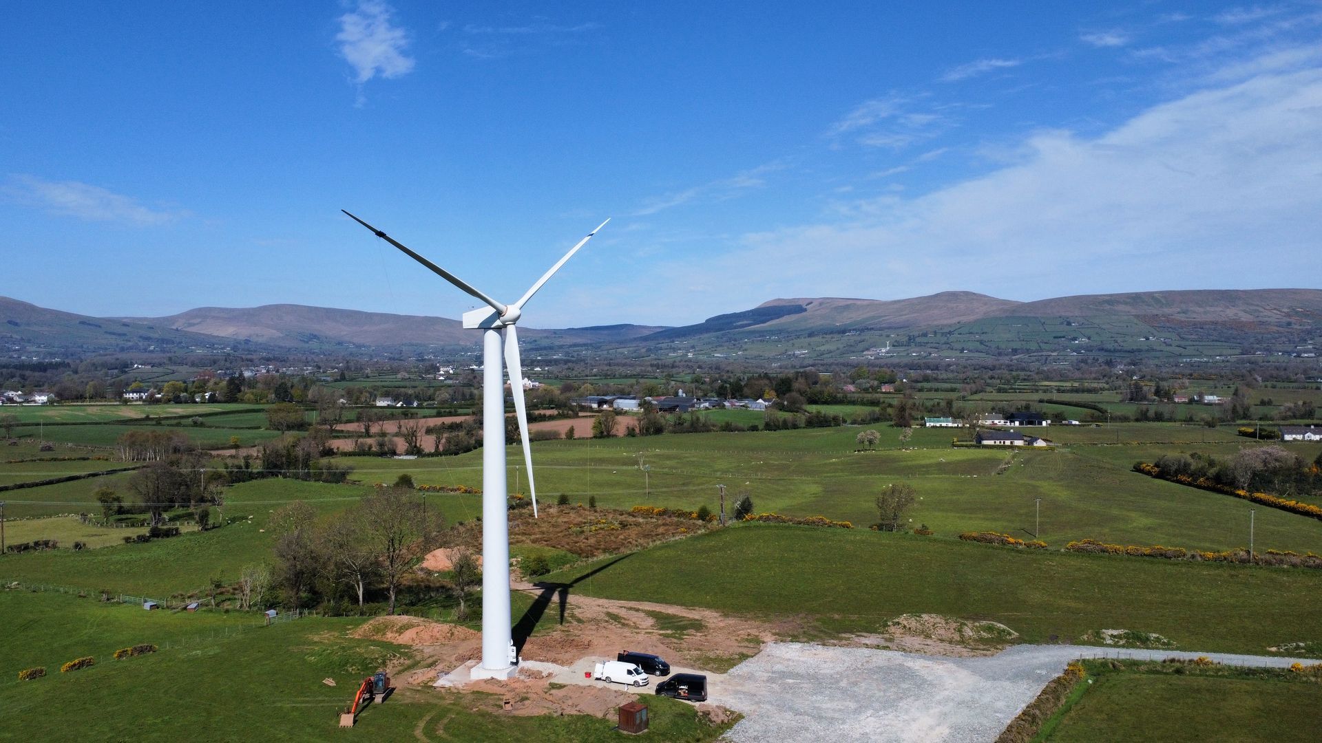 White wind turbine in a green rural field under a blue sky with mountains in the background